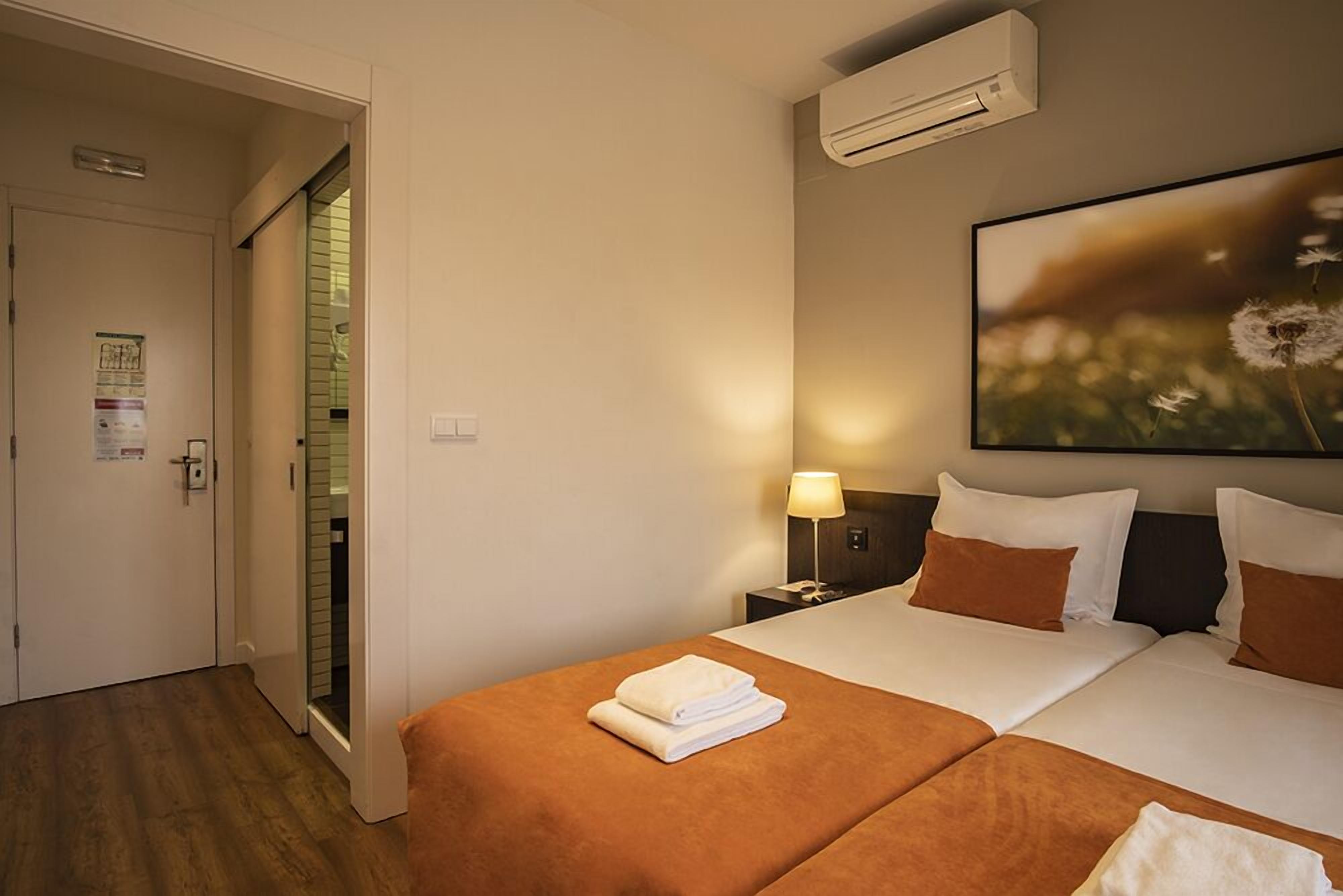Una habitación de hotel con cama, mesita de noche, lámpara y aire acondicionado instalado en la pared.