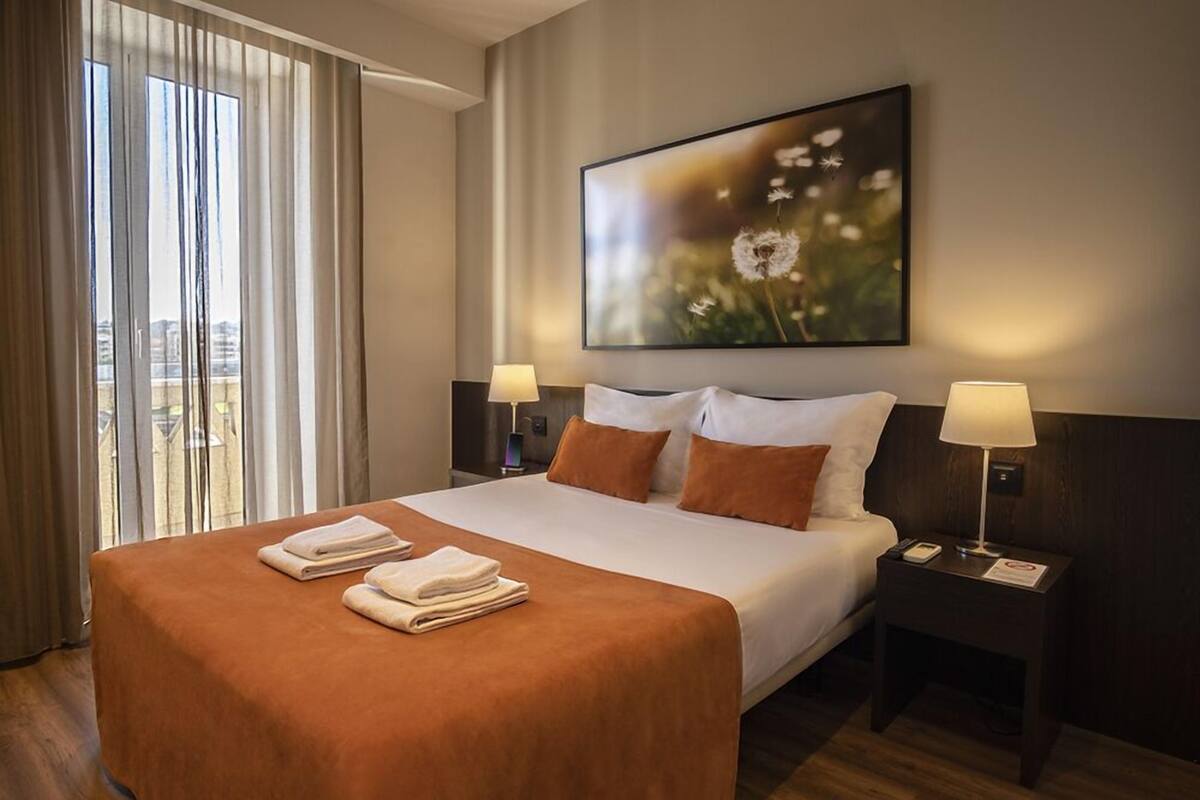 Habitación de hotel con una cama grande, dos lámparas de noche, un cuadro de diente de león y vistas a través de la ventana.