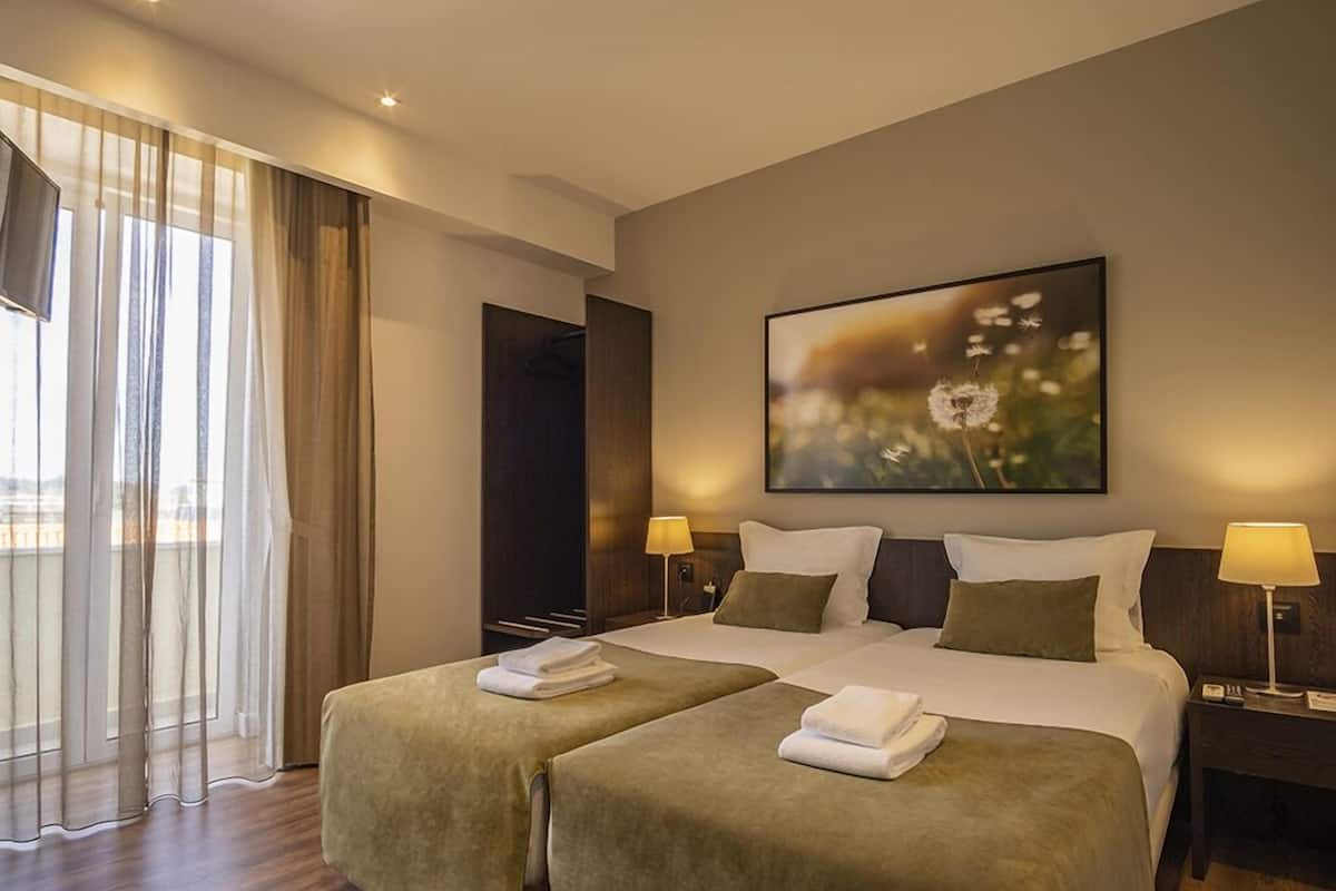Habitación de hotel con dos camas, un ventanal con cortinas translúcidas, un cuadro en la pared y lámparas de noche.