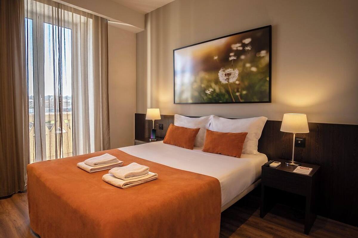 Habitación de hotel con una cama grande, dos lámparas de noche, un cuadro de diente de león y vistas a través de la ventana.