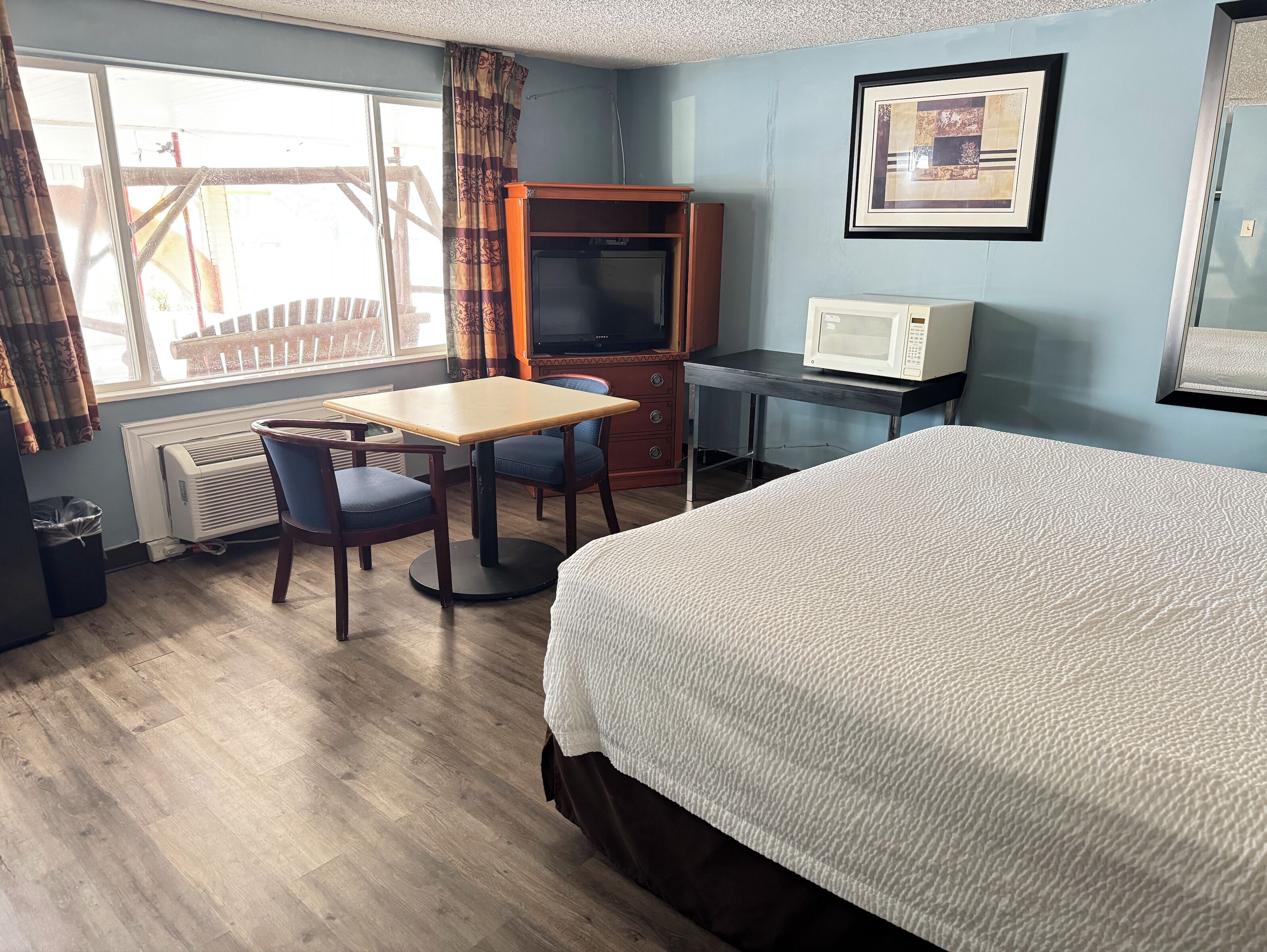 A hotel room with a bed, a desk with a microwave, a television, a window with curtains, and a radiator.