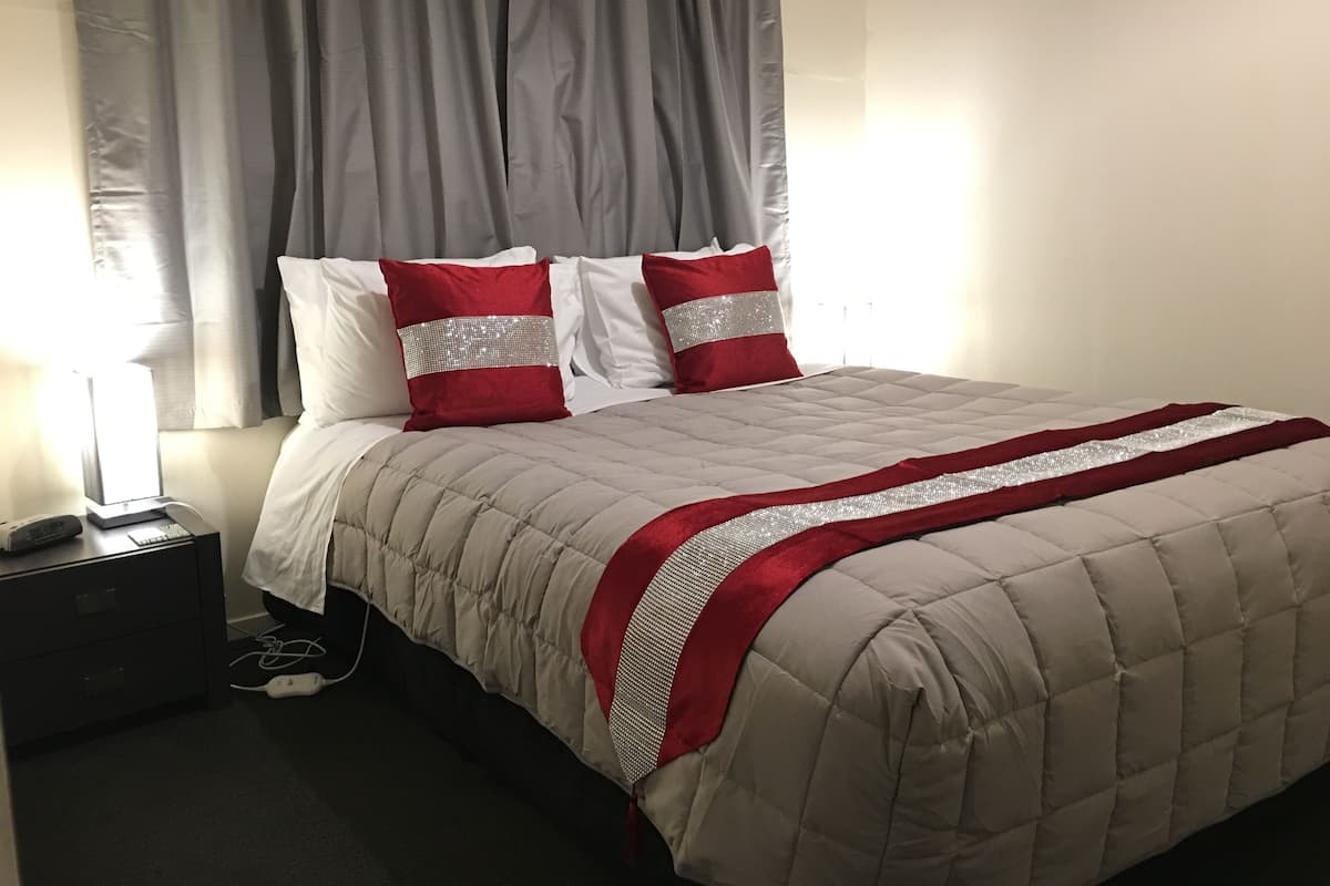 Uma cama bem arrumada com cobre acolchoado e almofadas vermelhas e brancas, um abajur e uma régua de tomadas em uma mesinha de cabeceira escura.