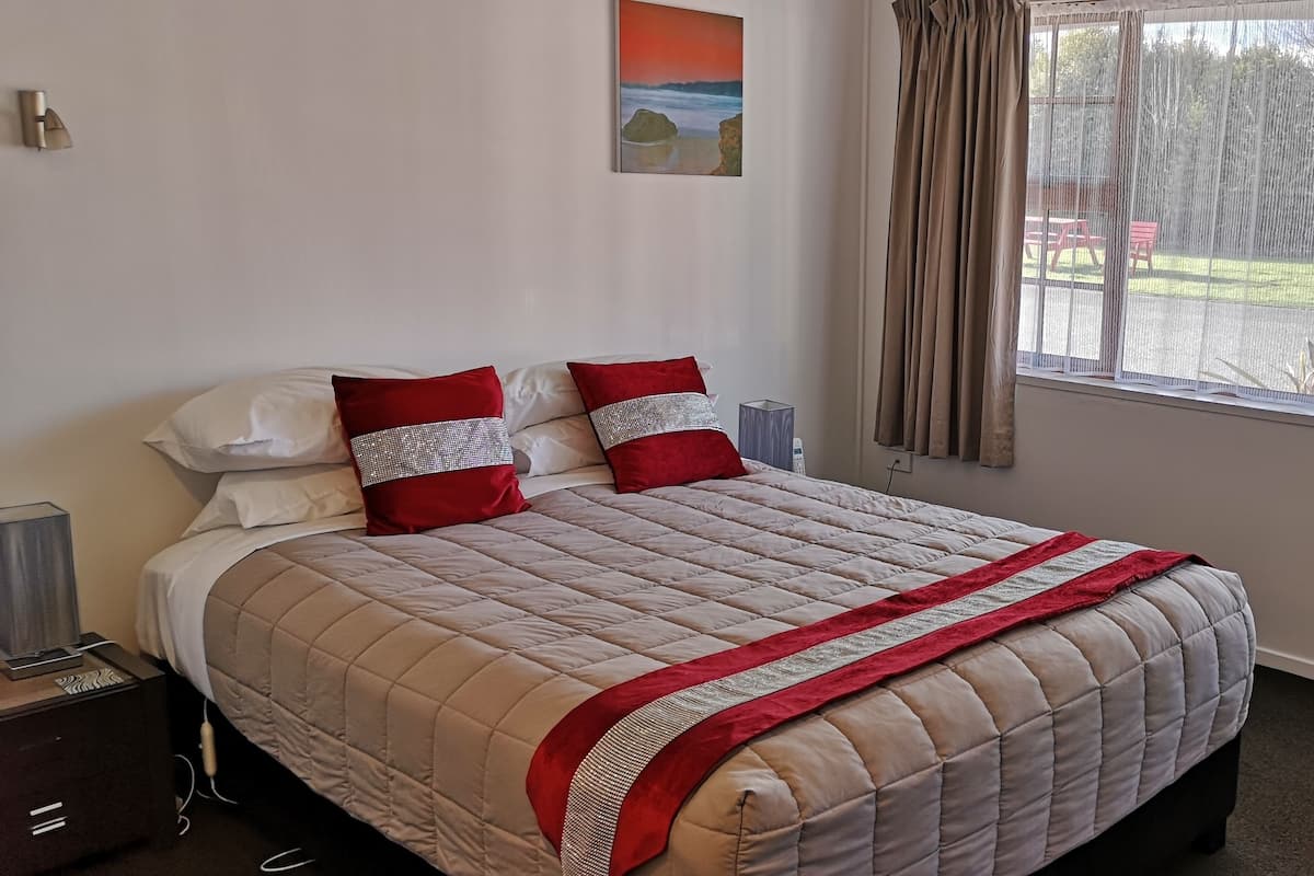 Cama bem arrumada com colcha bege e detalhes vermelhos, mesa de cabeceira com abajur, quadro na parede e janela com cortinas.