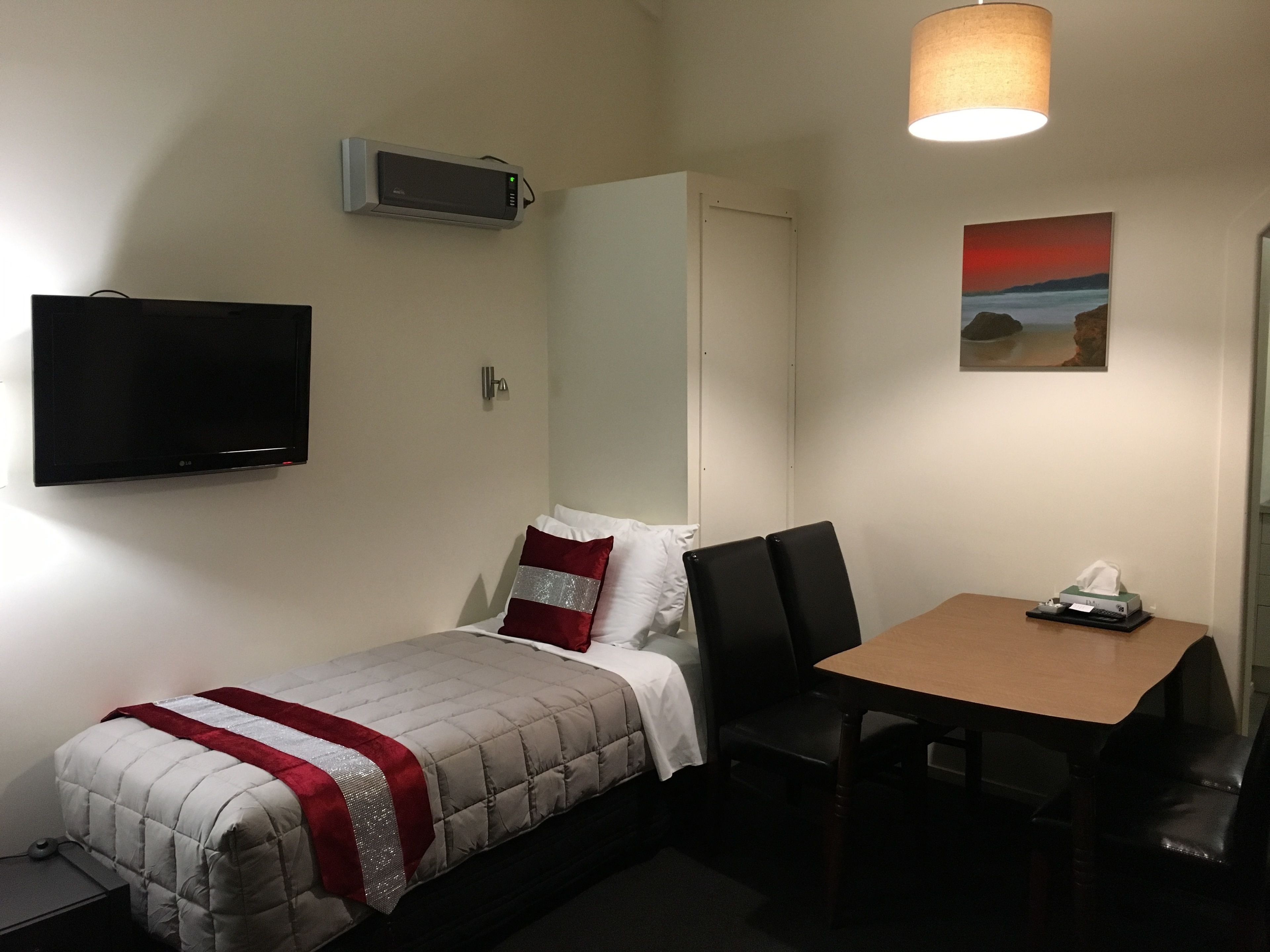 A small, neatly arranged room with a bed, a desk, a chair, a television, and a picture on the wall.