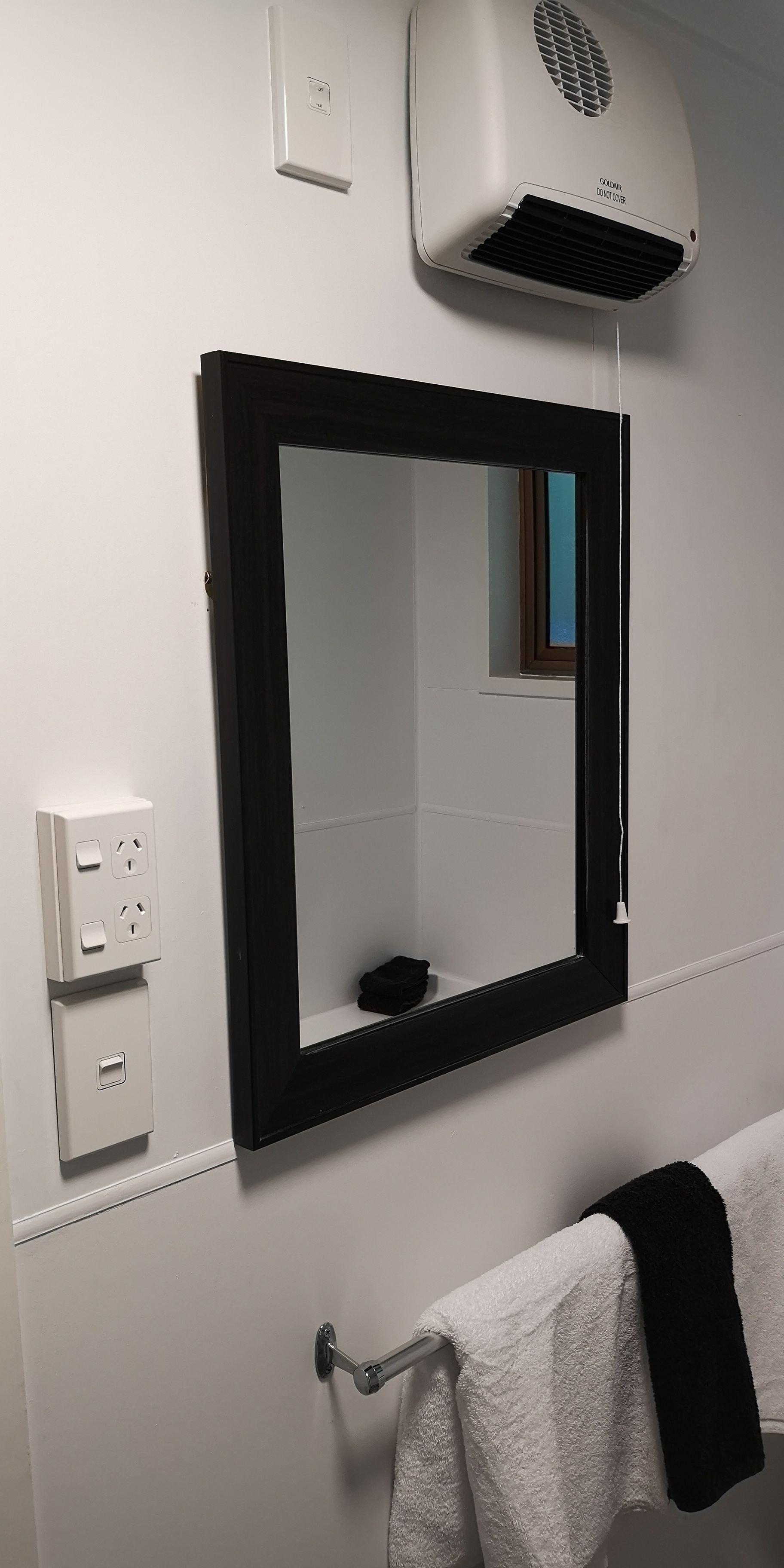 Un baño con espejo de marco negro, un perchero con toalla blanca, un secador de pelo y una ventana con vistas.