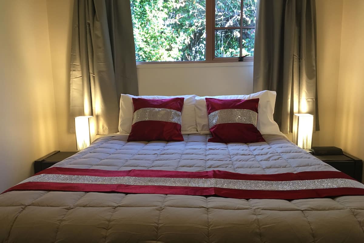 Uma cama bem arrumada com cobertura acolchoada e duas almofadas, uma mesa de cabeceira com abajur, e uma janela com cortinas.