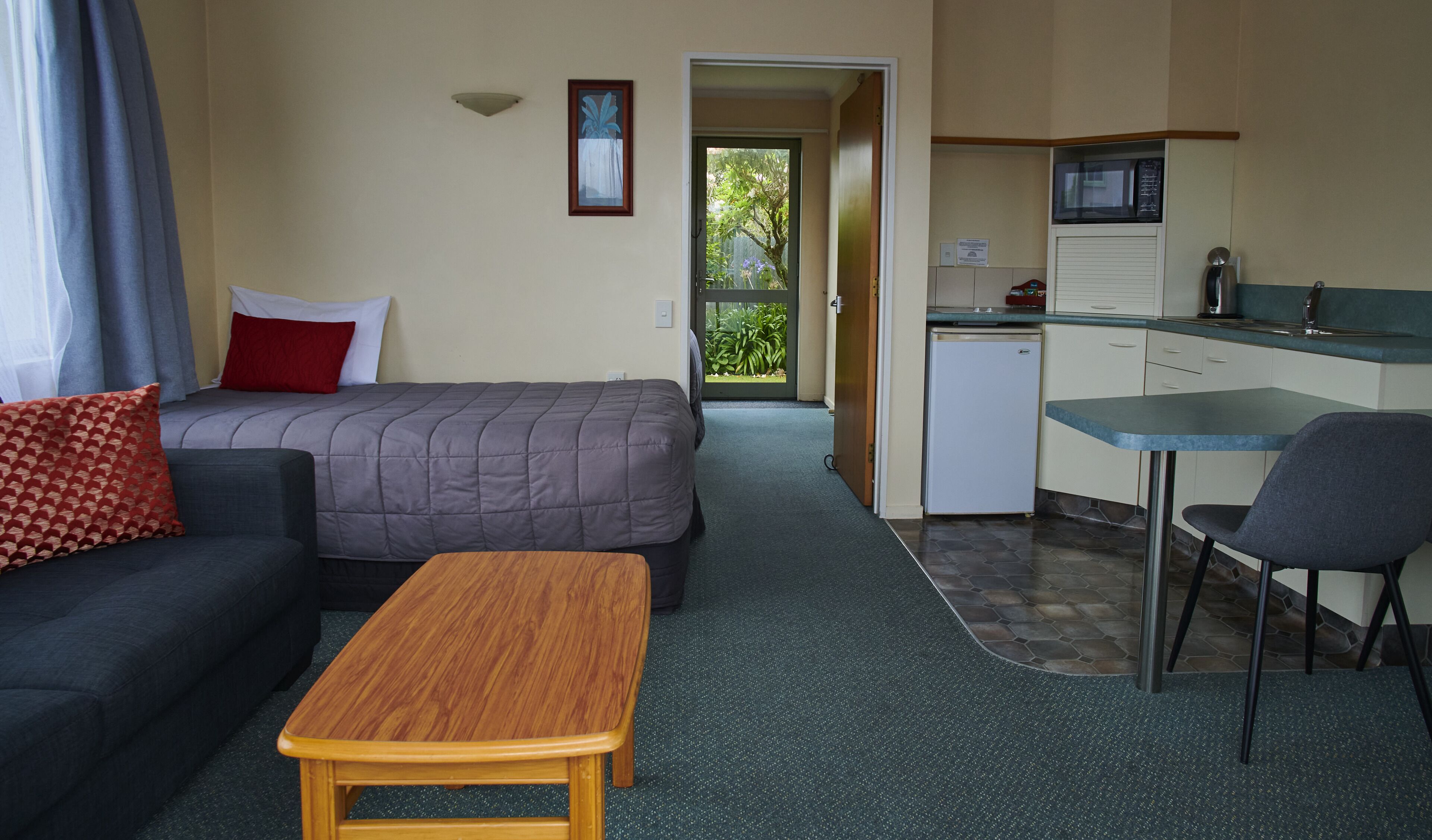 A compact hotel room with a bed, a sofa, a small table, a kitchenette, and a view of the outdoors.