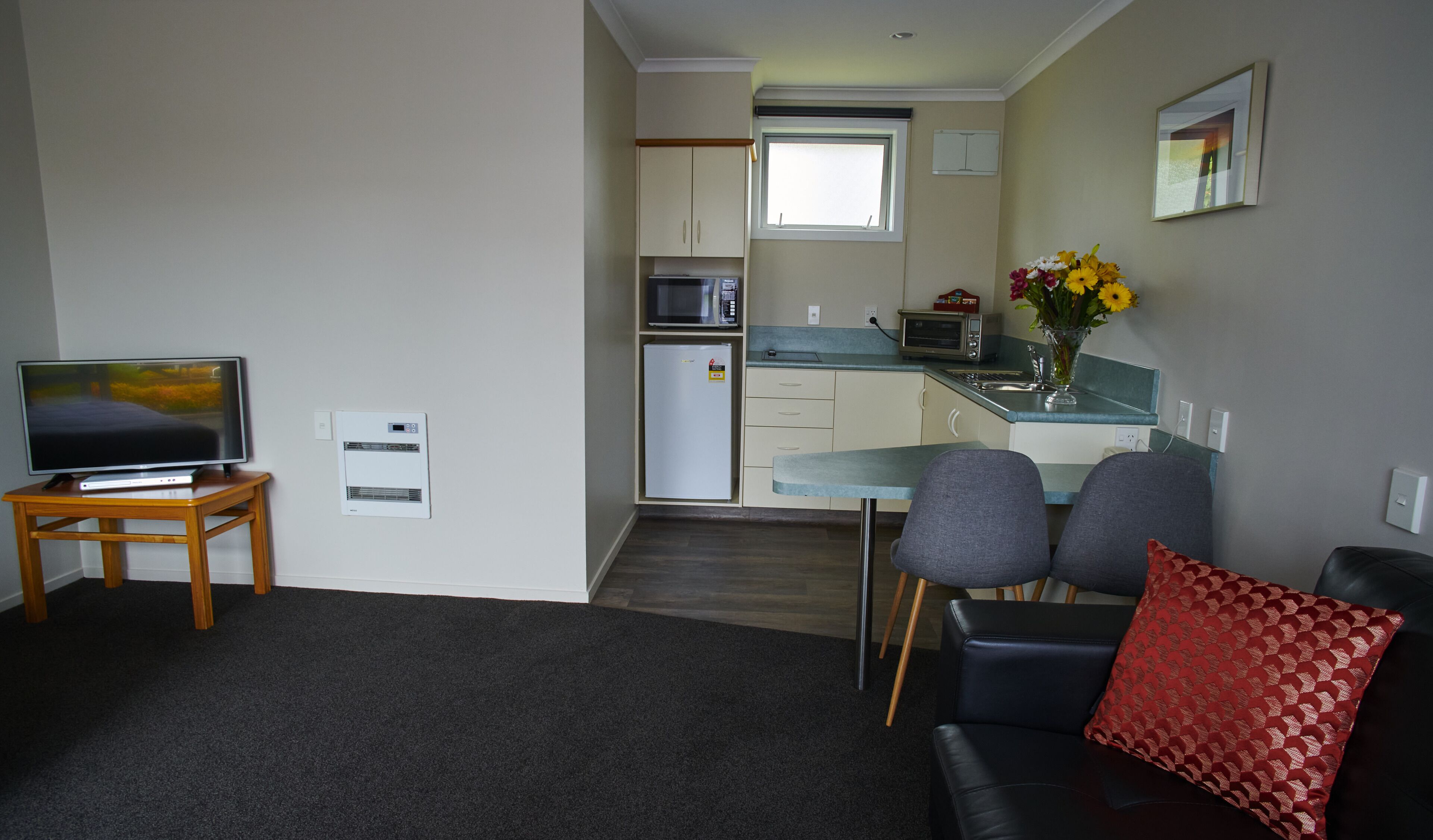 A compact living space with a television on a wooden stand, a kitchenette with a microwave, refrigerator, and toaster, and a dining area with two chairs and a red patterned pillow.
