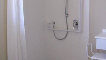 A small, white bathroom with a shower, a white chair, and a white shelf.