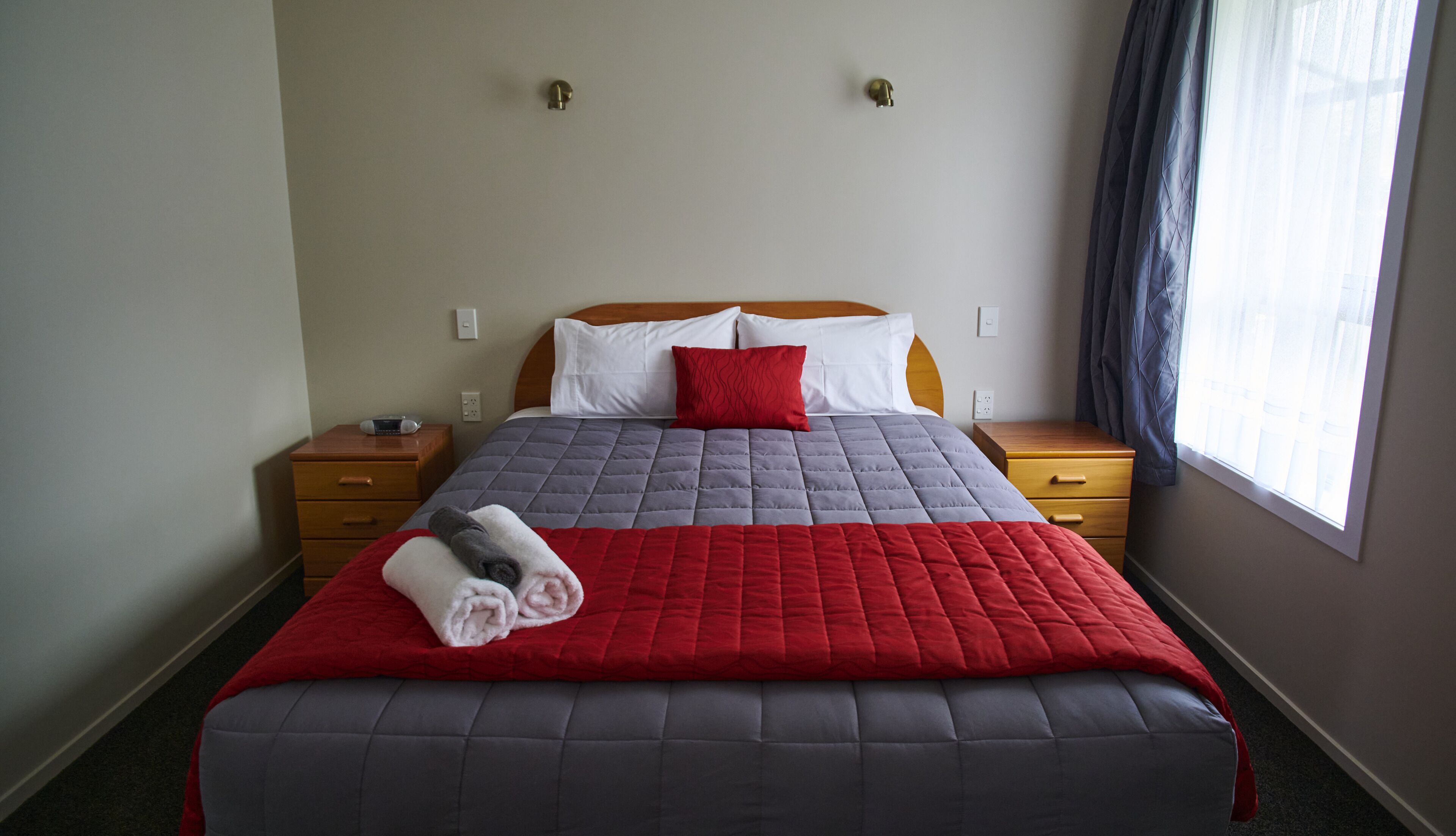 A hotel room with a bed, two nightstands, a red and grey quilted bedspread, a red pillow, and a window with curtains.