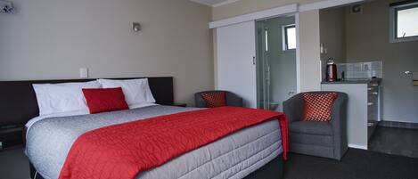 A modern hotel room with a large bed, a gray armchair, a kitchenette, and a bathroom with a glass shower.