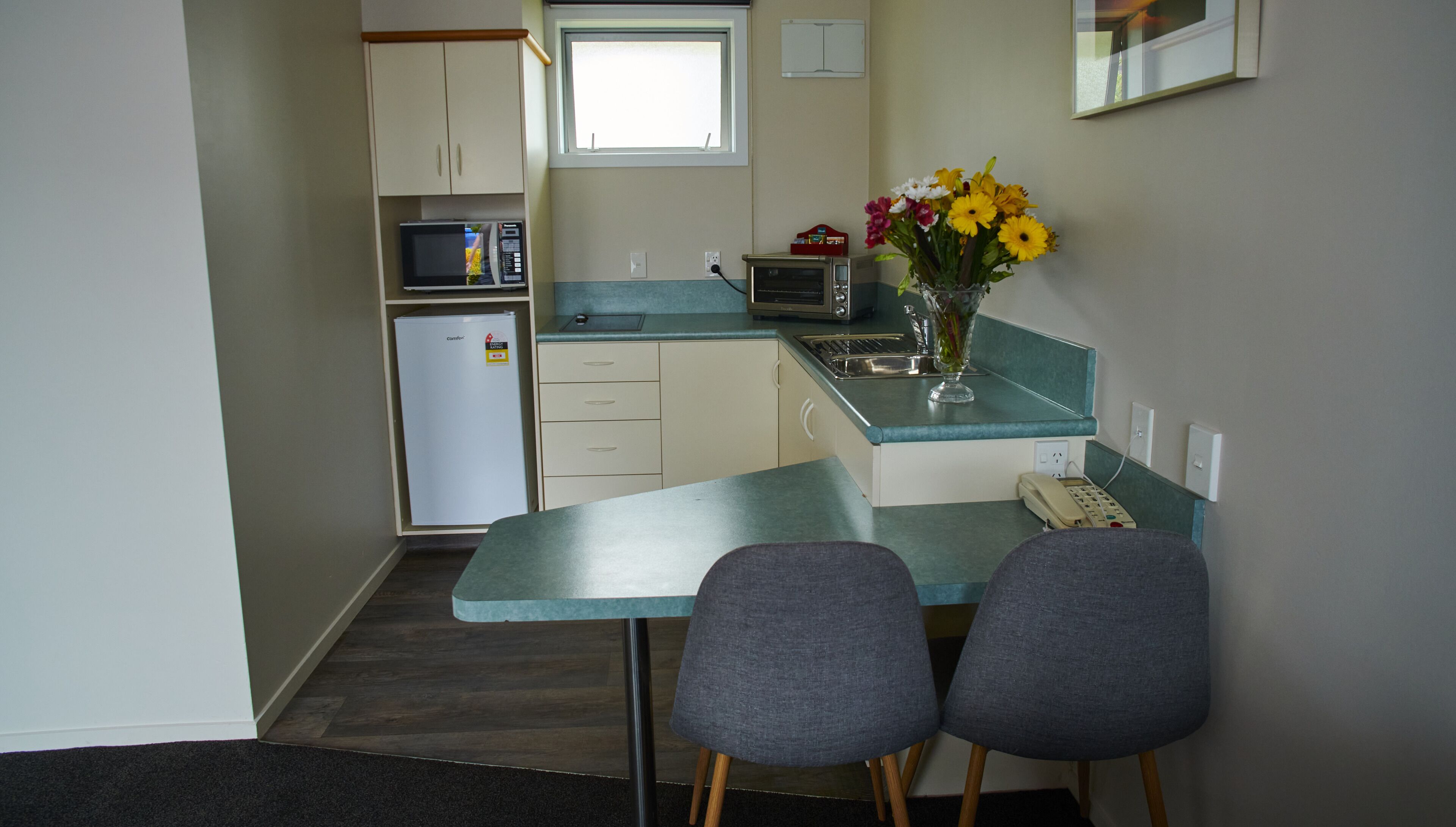 A compact kitchen with a microwave, toaster, and refrigerator. There are two chairs at a table, and a vase with flowers on the counter.