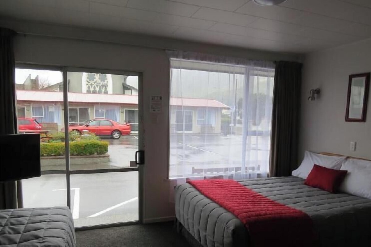A hotel room with a bed, a television, and a window with a view of a parking lot and buildings.