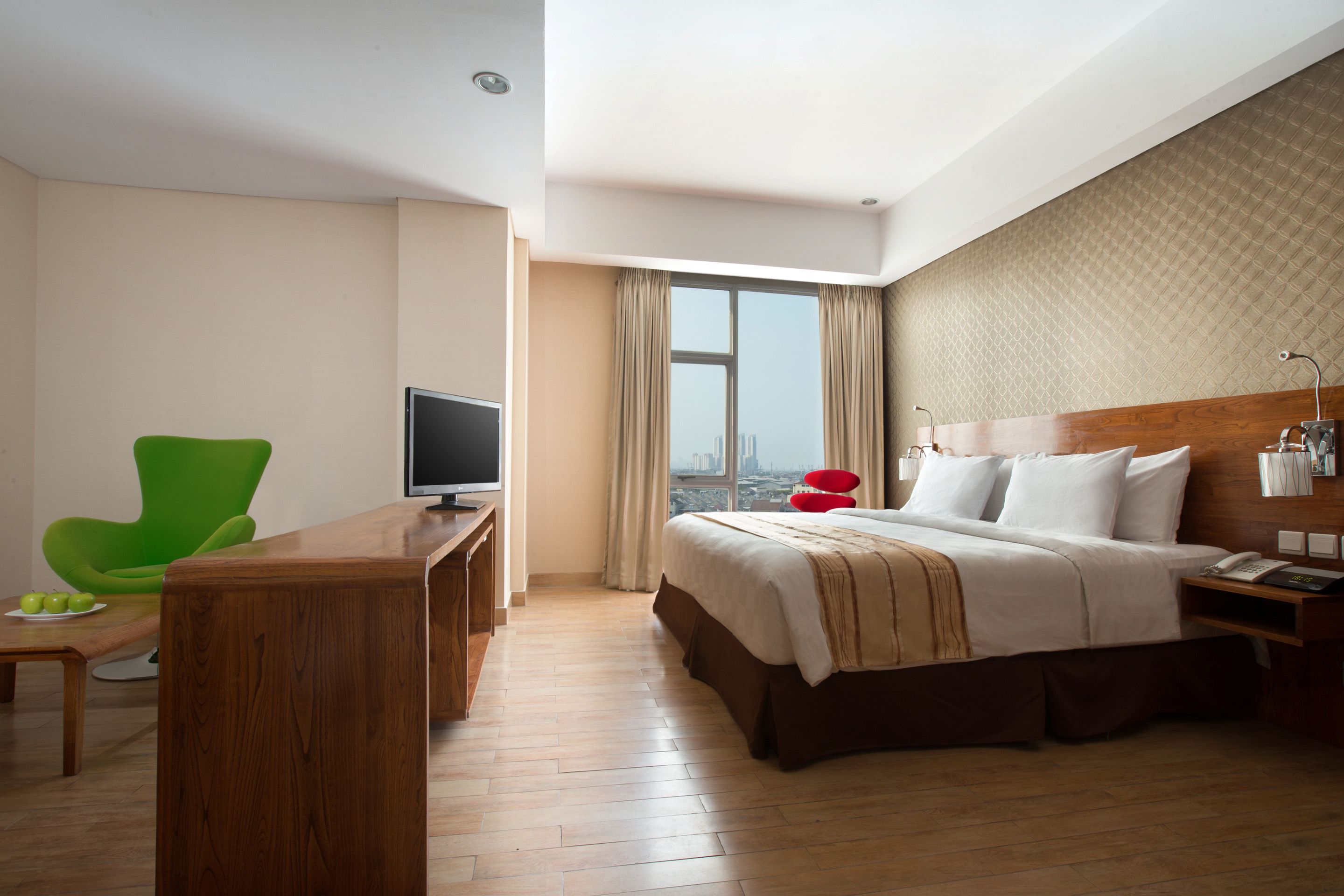 Ein Hotelzimmer mit einem Bett, einem Schreibtisch mit Fernseher, einem grünen Sessel und einem großen Fenster mit Blick auf die Stadt.