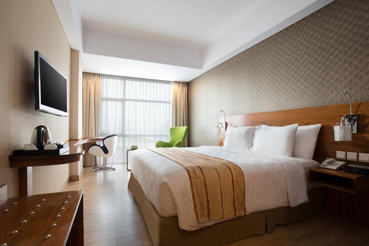 A hotel room with a large bed, a TV mounted on the wall, a desk with a telephone, a green chair, and a window with curtains.