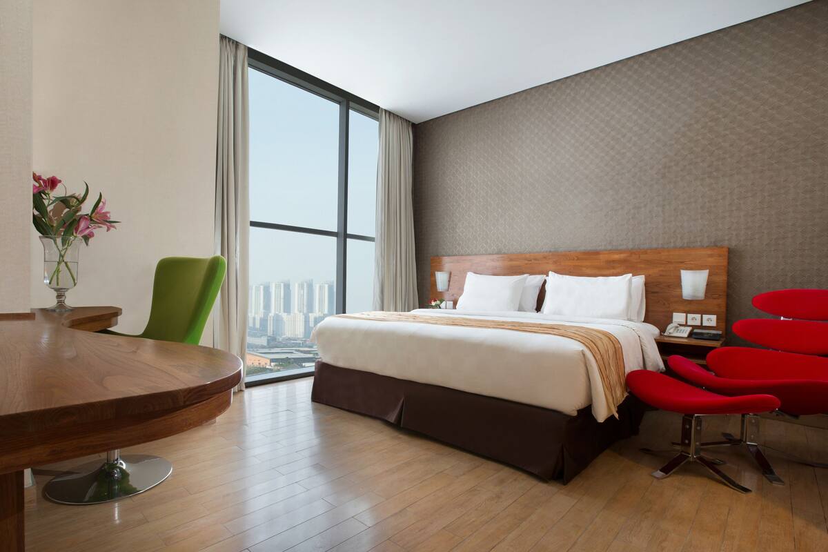 A modern hotel room with a large bed, a wooden desk, a green chair, and a view of the city.