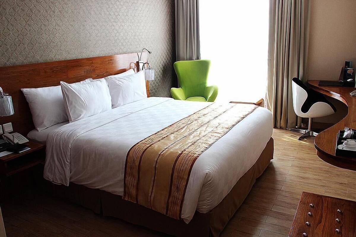 A hotel room with a large bed, a green armchair, a desk with a lamp, and a wooden nightstand with a phone.
