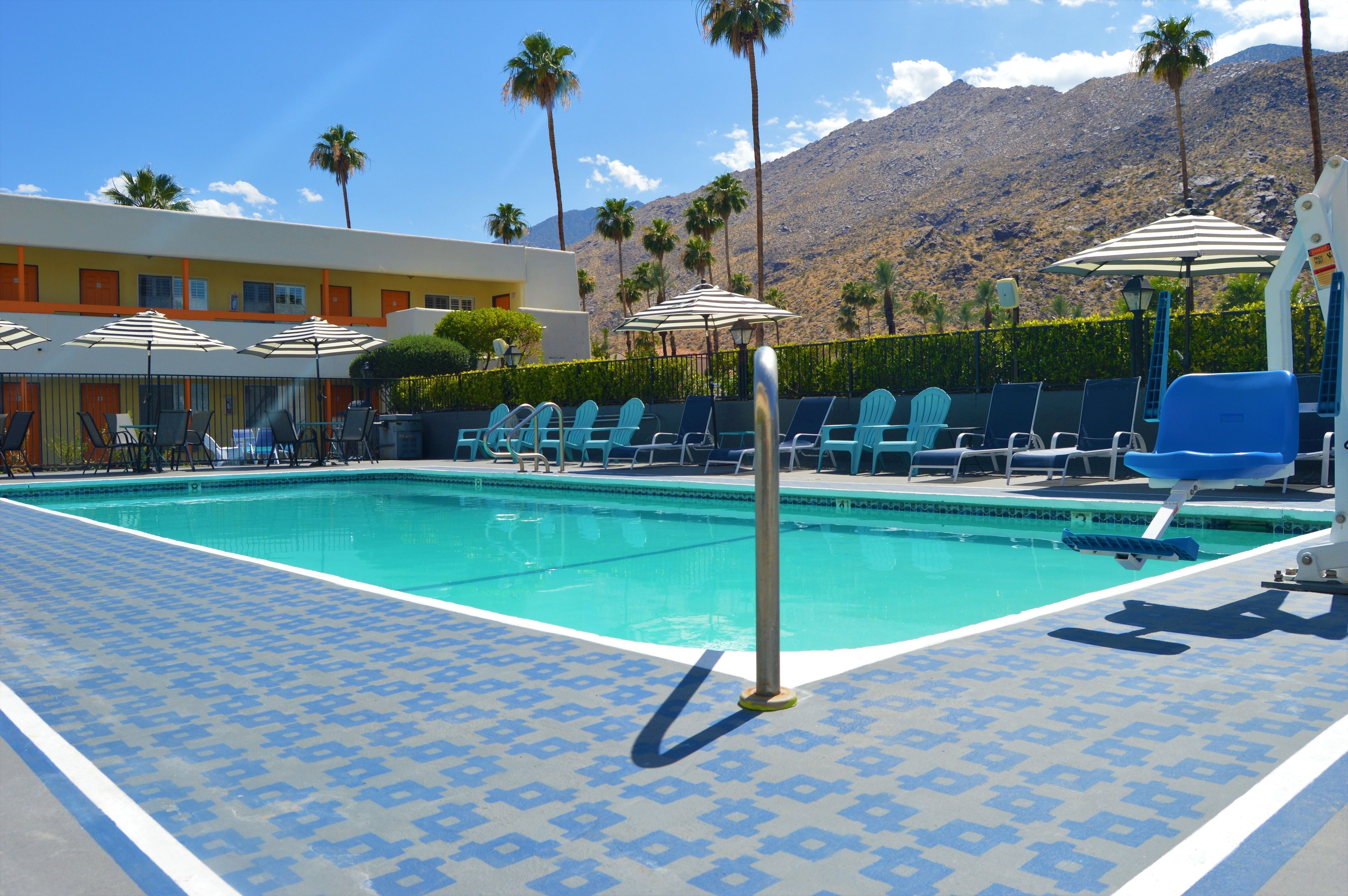 Area piscina con sedie a sdraio e ombrelloni, con montagne sullo sfondo e un edificio con porte arancioni.