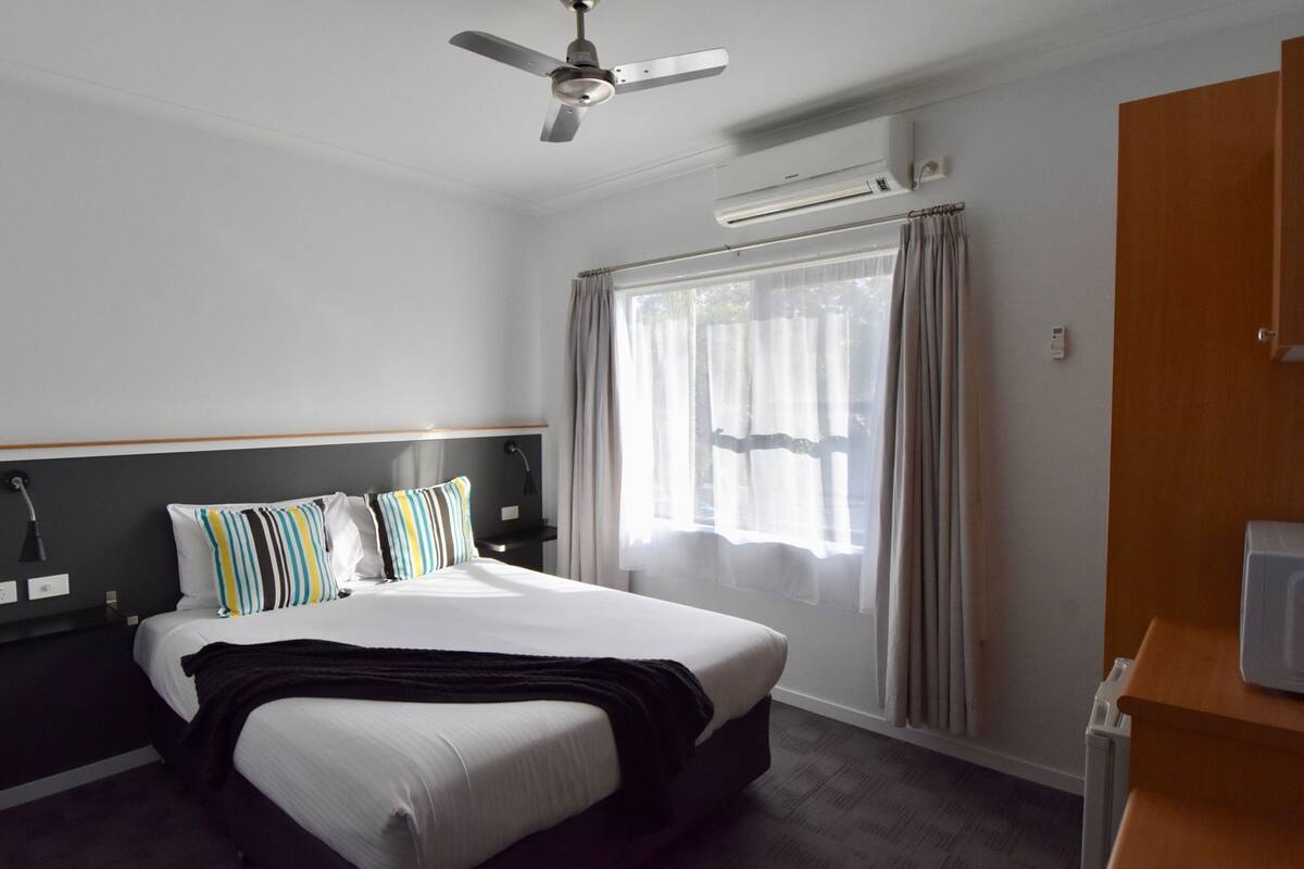 A hotel room with a bed, a ceiling fan, an air conditioning unit, a window with curtains, and a wooden desk.