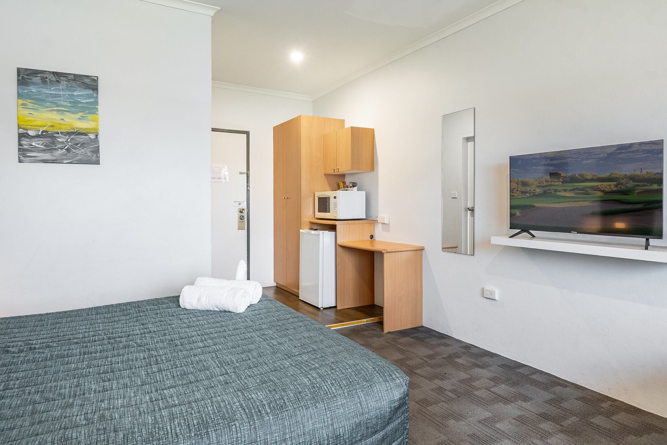 A hotel room with a bed, a kitchenette, a flat-screen TV, and a painting on the wall.