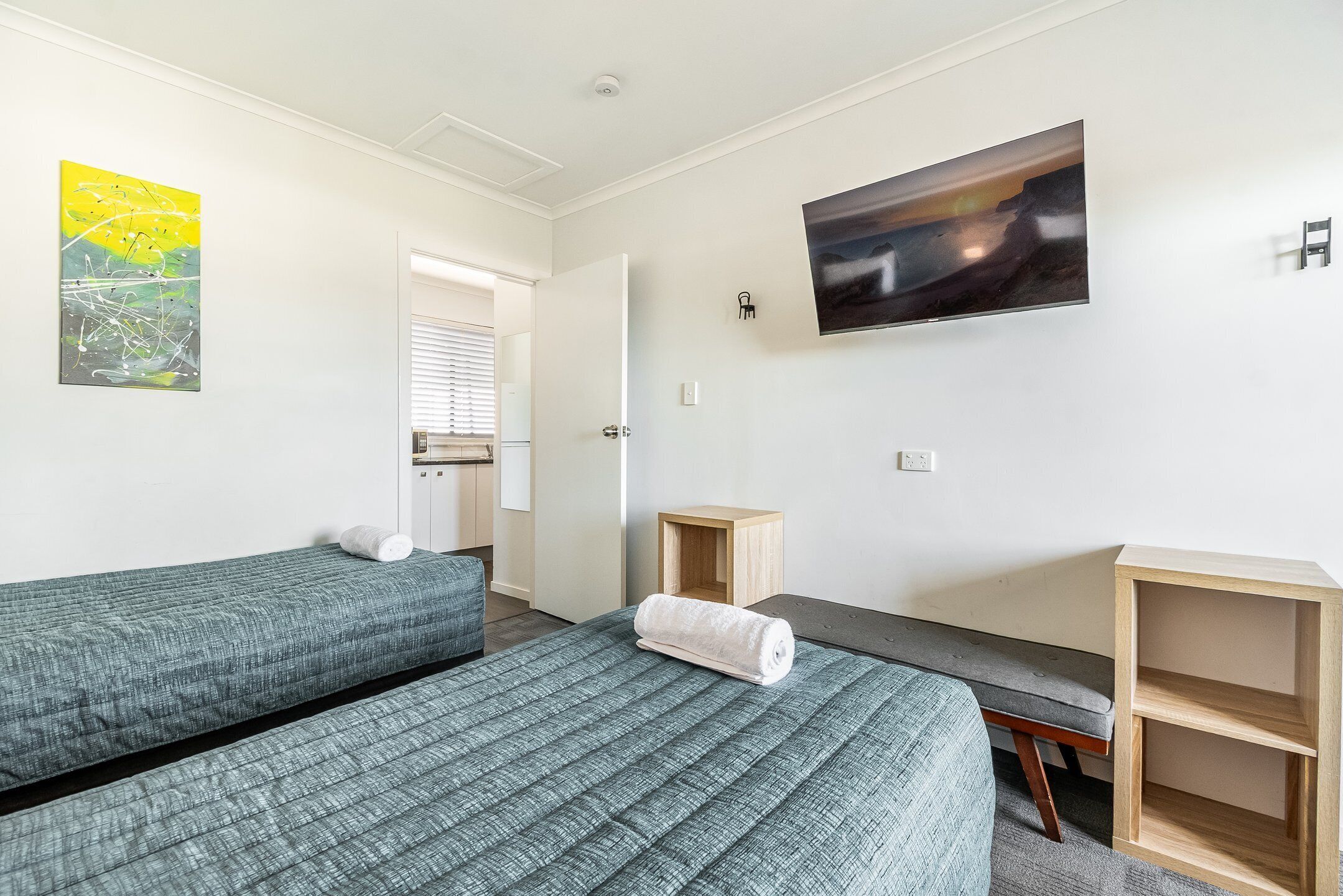 Habitación de hotel con dos camas individuales, un pequeño escritorio de madera, televisor montado en la pared y una ventana con persianas.