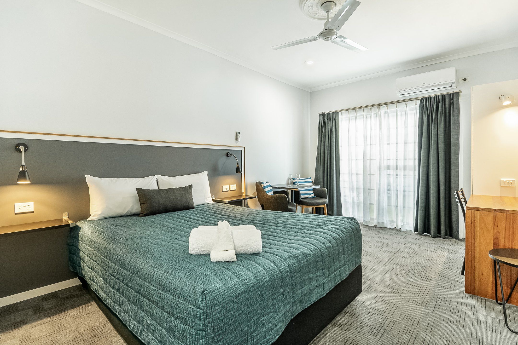 Habitación de hotel con cama, mesitas de noche, escritorio, silla, ventilador de techo y ventana con cortinas.