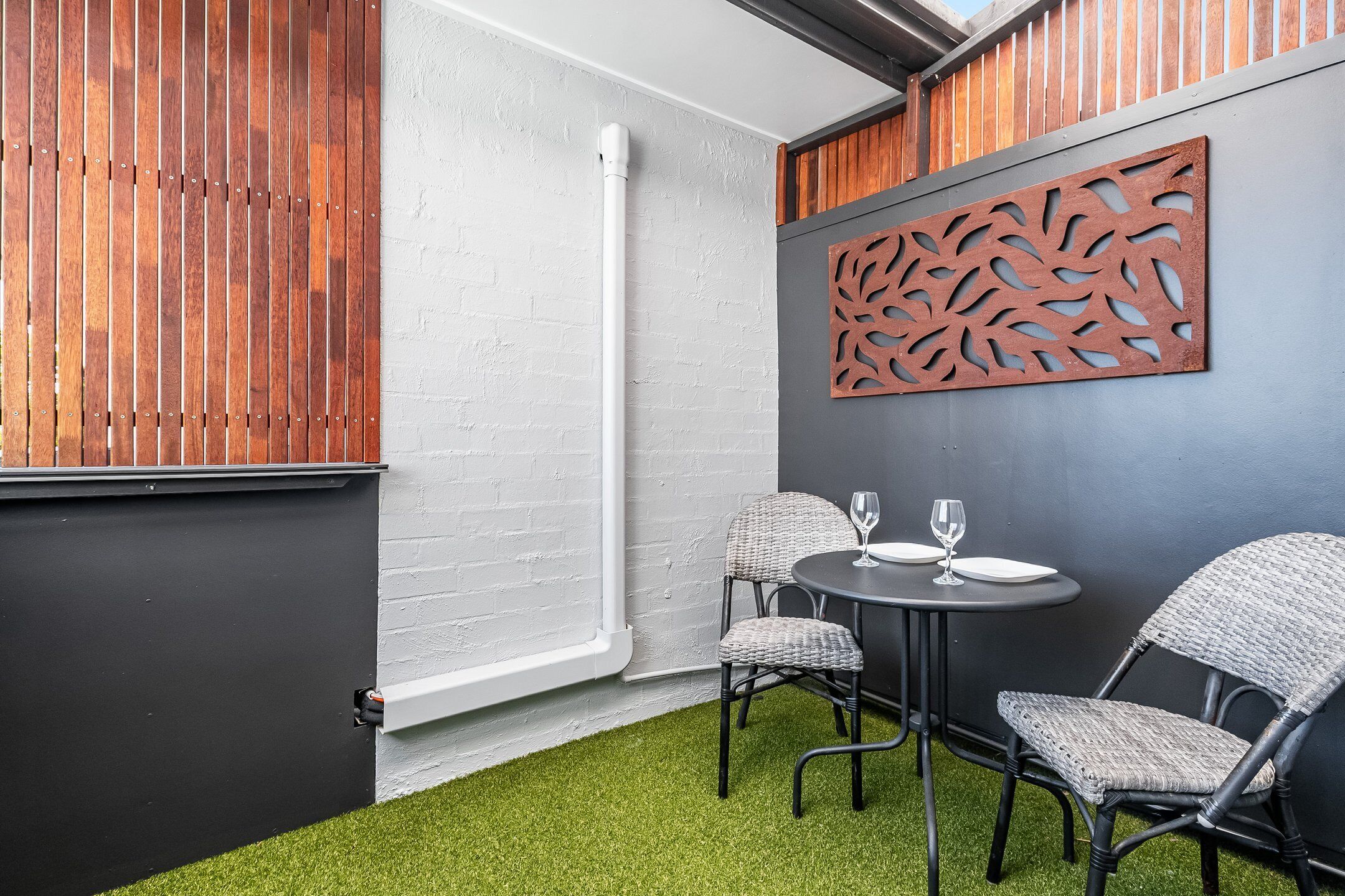 A corner room with a black door, white brick wall, and a decorative panel. There is a round table with two chairs and a glass on it.