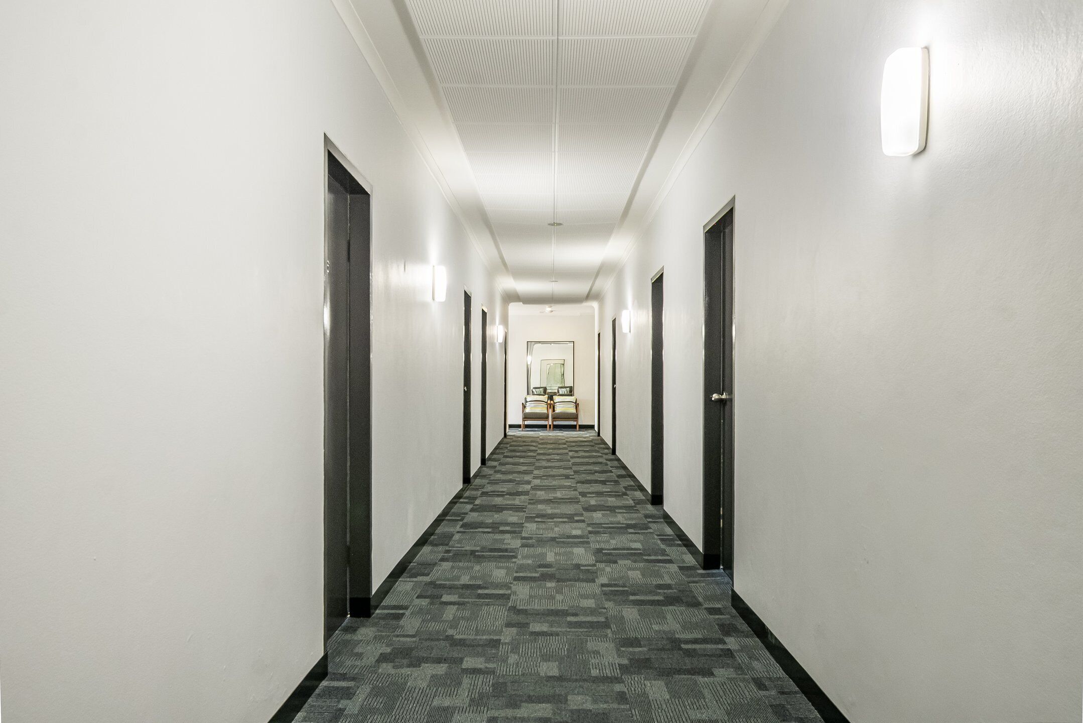 A hotel hallway with a carpeted floor, white walls, and doors on both sides.