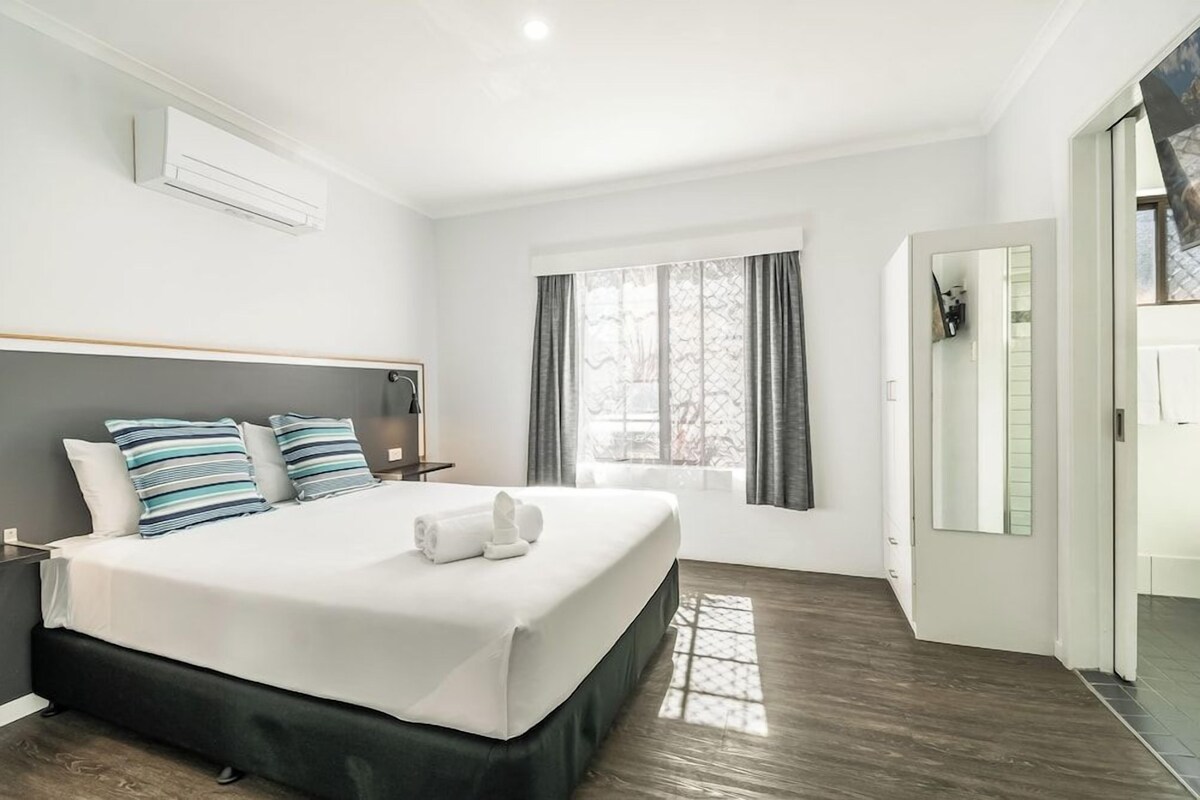 A modern hotel room with a large bed, a bedside table, a window with curtains, and a door leading to a bathroom.