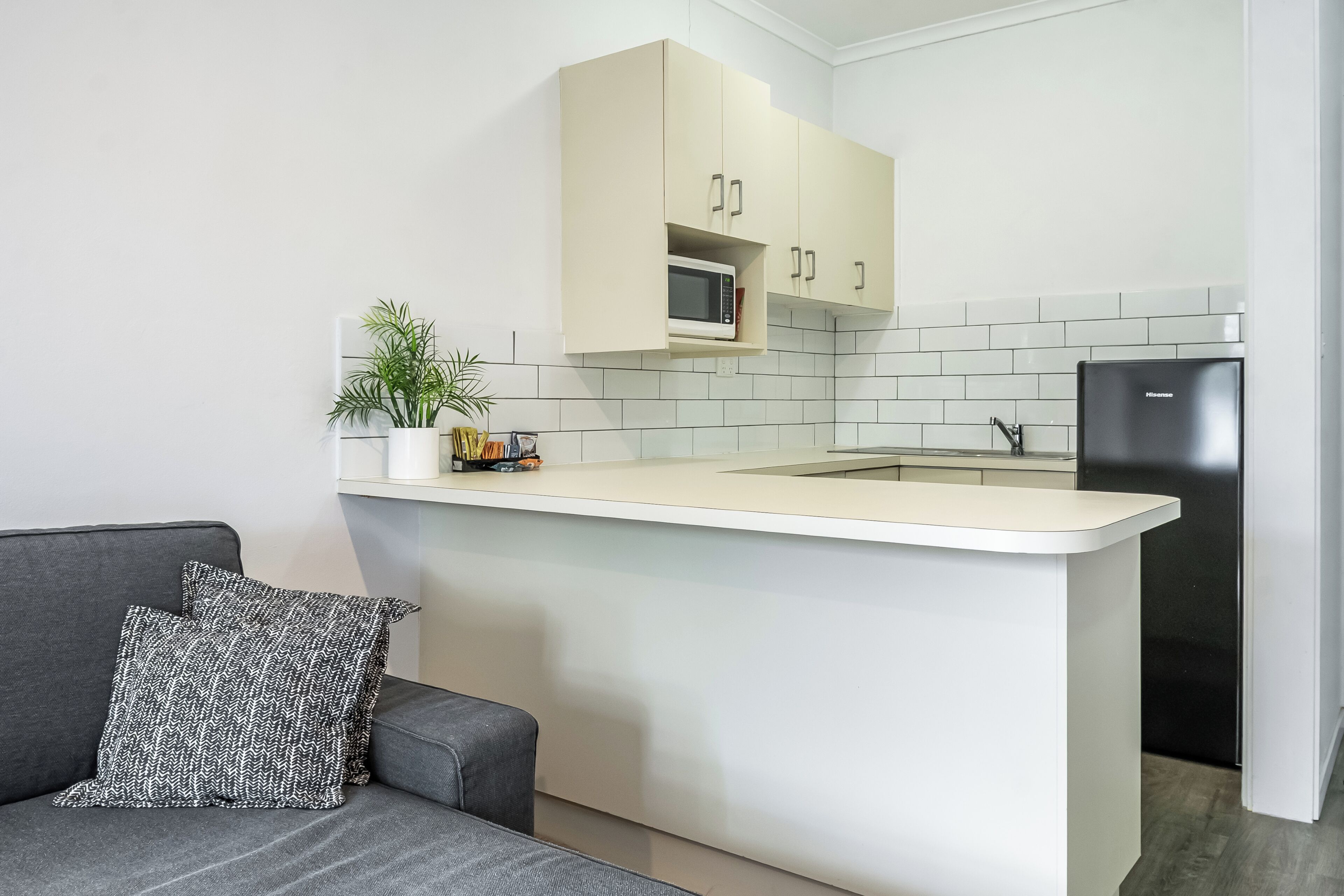 A compact kitchen with a white countertop, a microwave, a refrigerator, and a grey sofa with a patterned pillow.