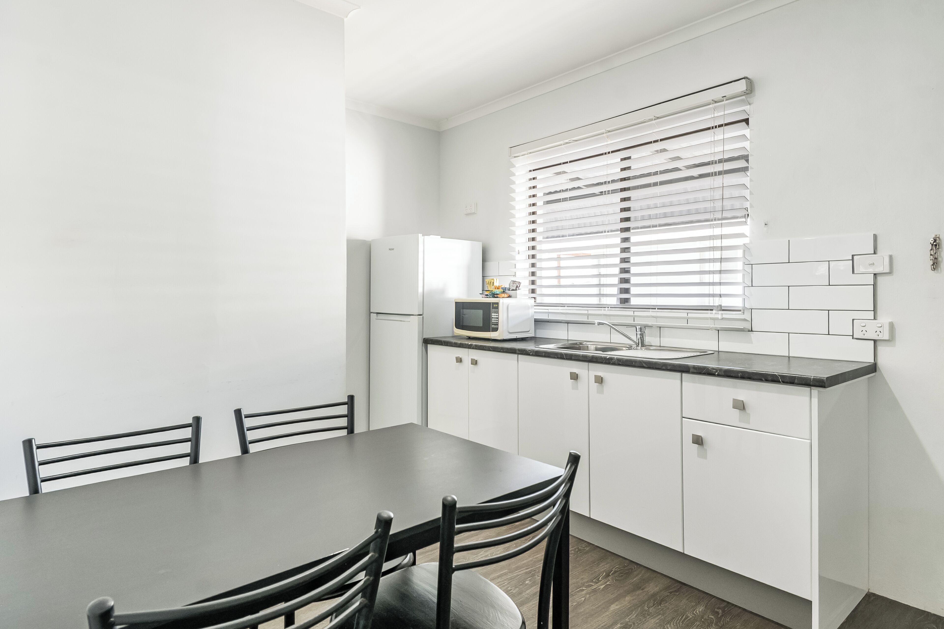A modern kitchen with white cabinets, a dark countertop, a microwave, and a window with blinds.