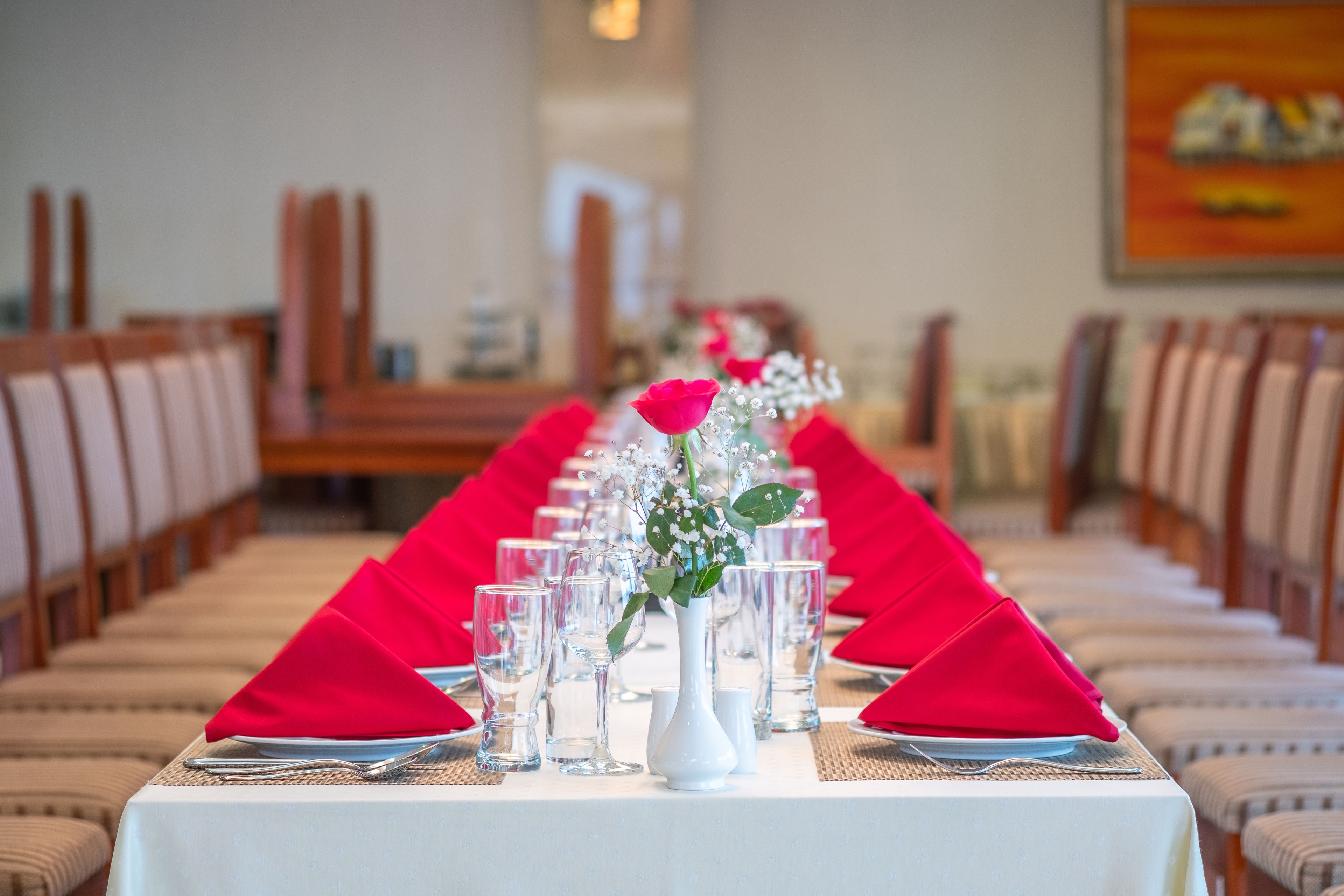 Mesa de jantar posta com guardanapos vermelhos, copos transparentes e um vaso branco com flores.