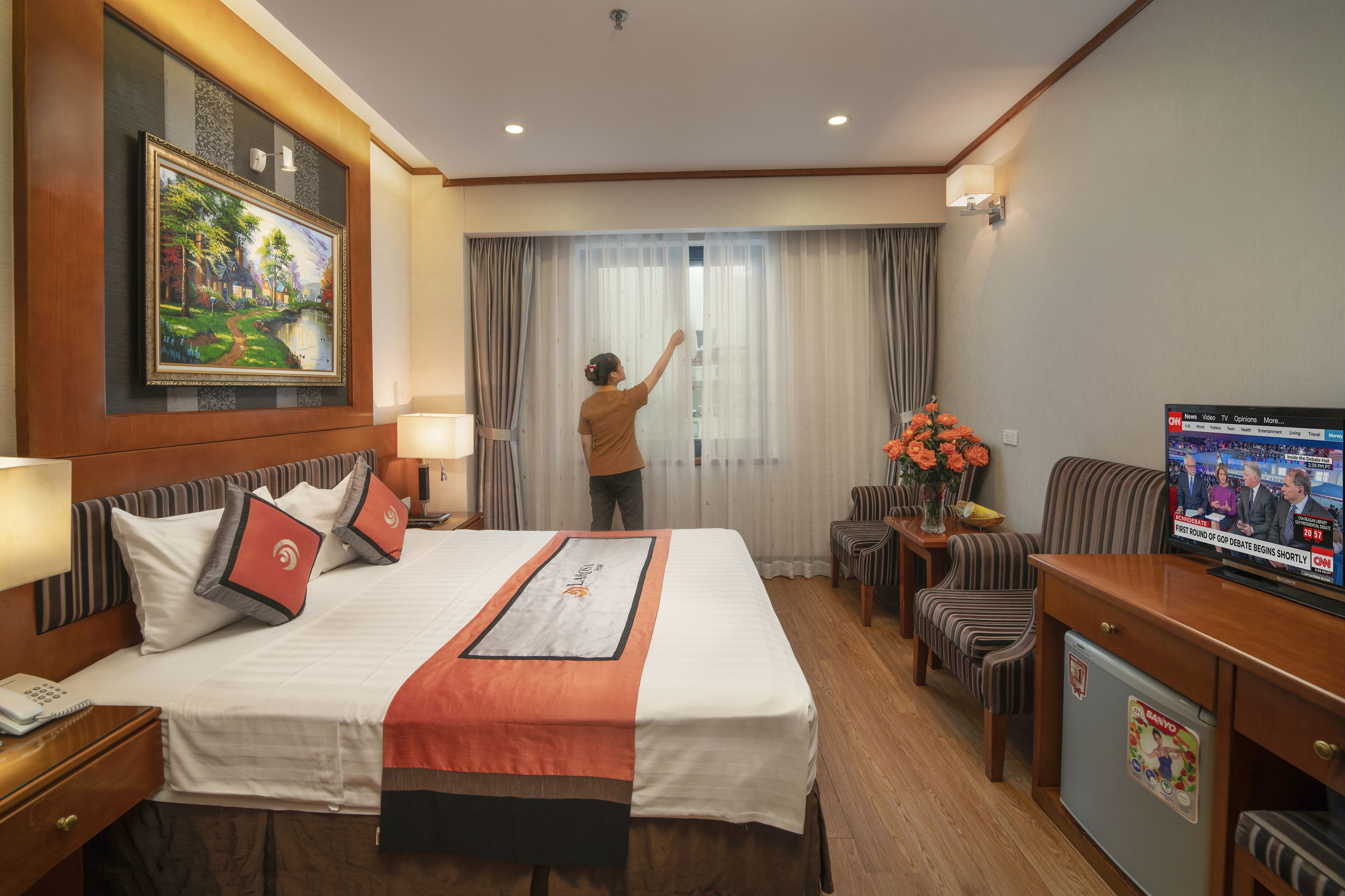 Una habitación de hotel con una cama bien hecha, televisión, escritorio con lámpara y un cuadro en la pared.
