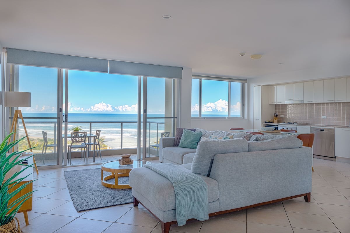 A spacious living room with a large sofa, a dining table, and a view of the beach.