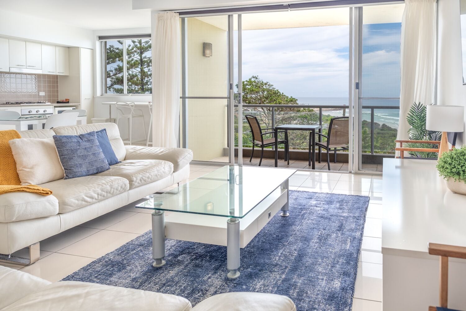 A modern living room with a glass coffee table, a sofa with cushions, and a view of the ocean through large sliding doors.