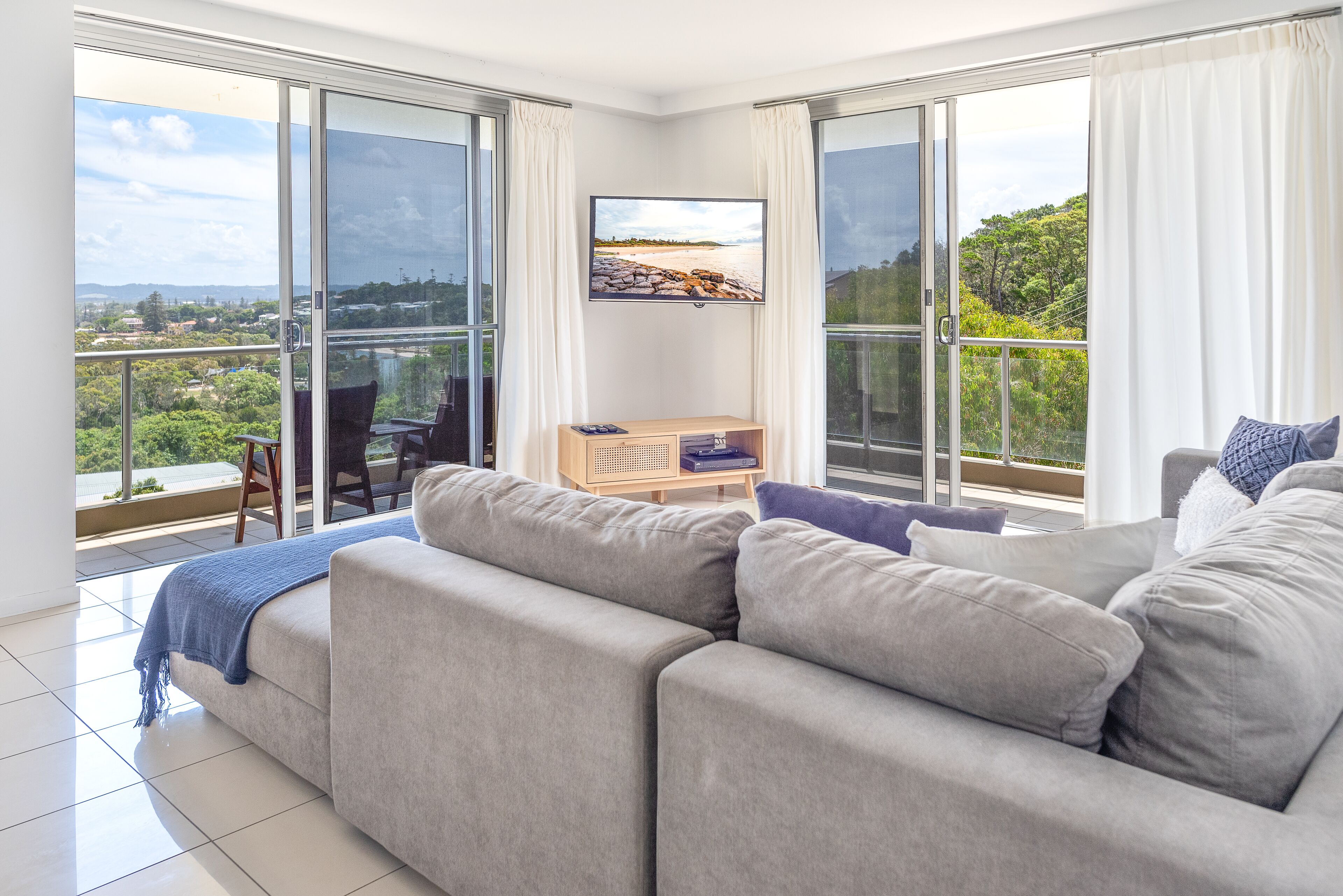A modern living room with a grey sofa, a television mounted on the wall, and a view of a cityscape through large windows.