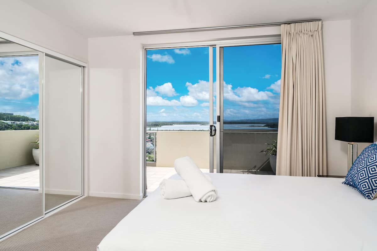 A hotel room with a large sliding glass door leading to a balcony, a bed with a rolled white towel, a black TV stand, and a patterned pillow.