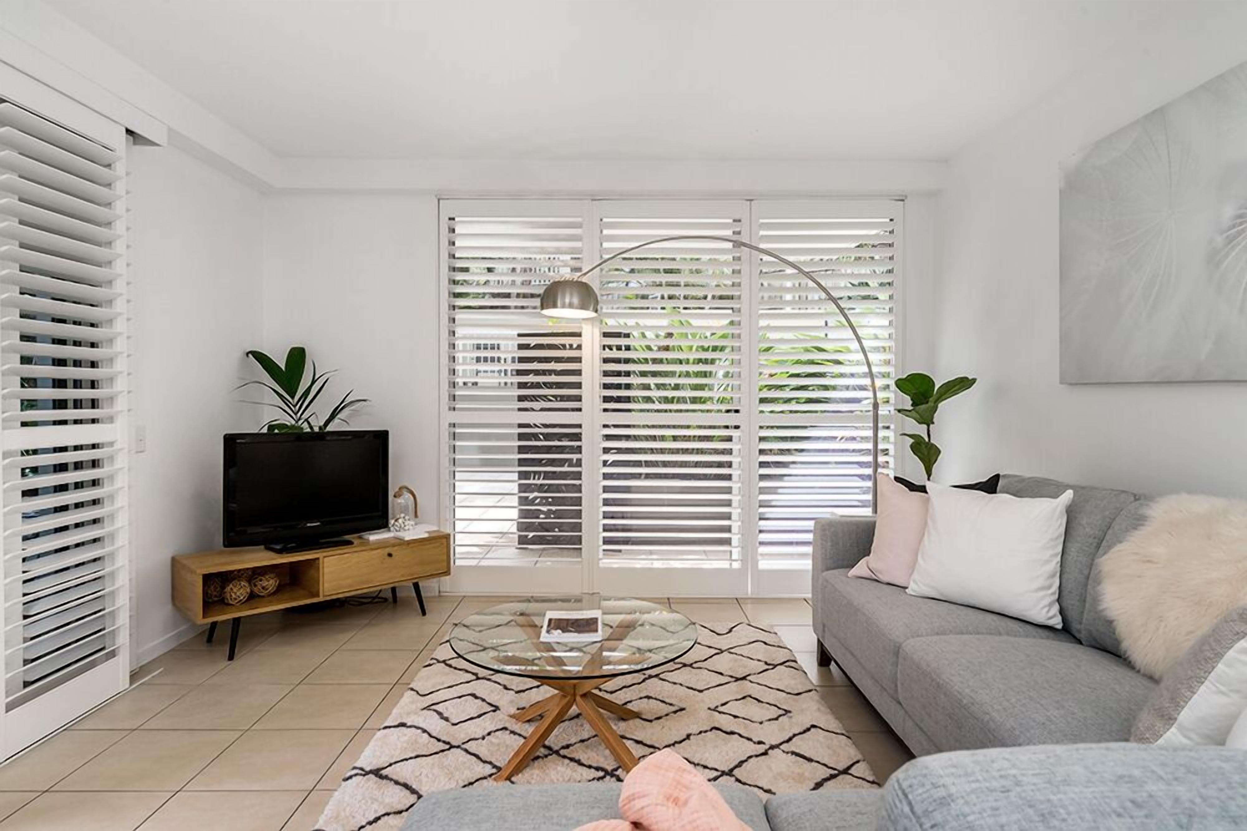 A living room with a sofa, a coffee table, a TV on a wooden stand, and a large window with blinds.