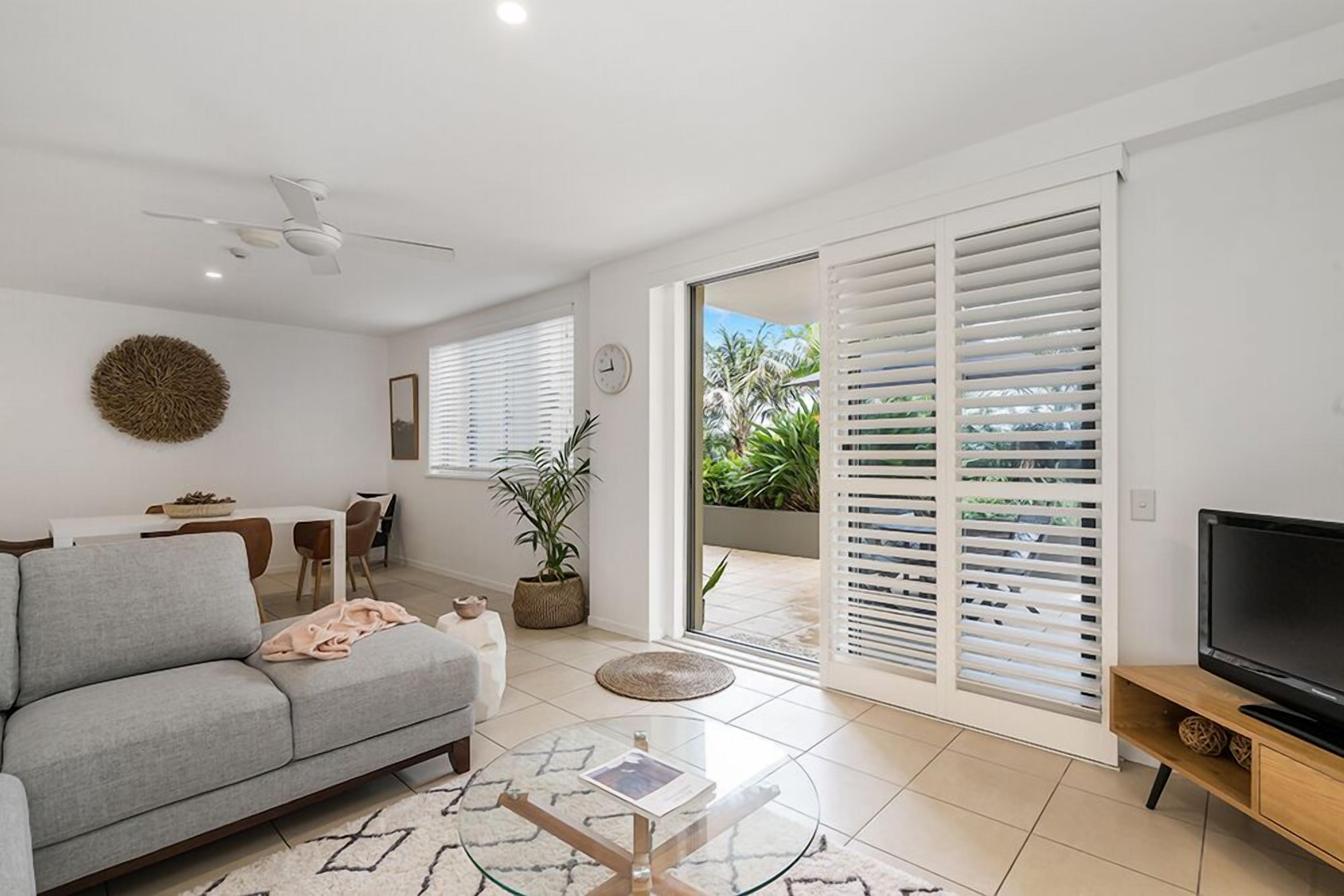 A modern living room with a grey sofa, a glass coffee table, a TV on a wooden stand, and a sliding glass door leading to a balcony with white blinds.