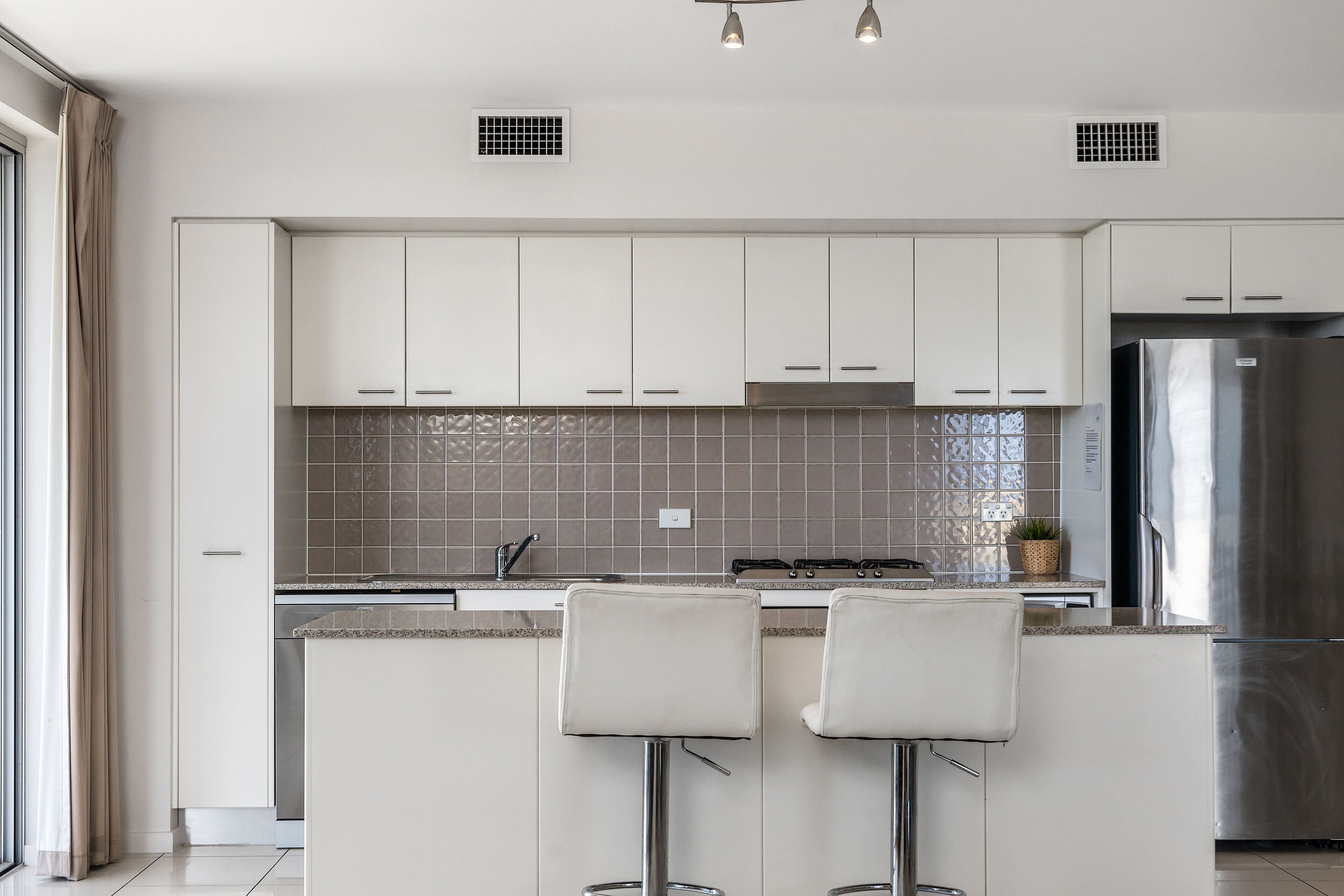 A modern kitchen with white cabinets, a stainless steel refrigerator, and two bar stools.