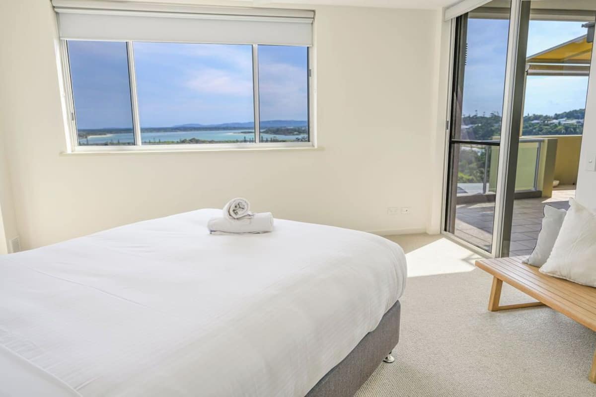 A bedroom with a large bed, a view of the sea through a window, and a balcony with a wooden bench.