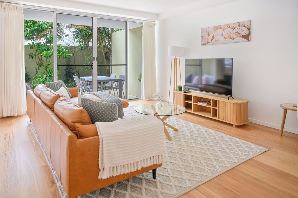 Un salon moderne comprenant un canapé, une table basse en verre, un téléviseur et une porte-fenêtre coulissante donnant sur un balcon aménagé avec du mobilier d’extérieur.