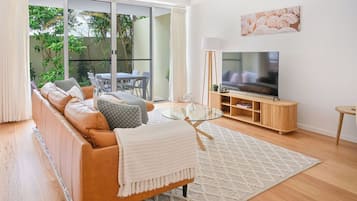Un salon moderne comprenant un canapé, une table basse en verre, un téléviseur et une porte-fenêtre coulissante donnant sur un balcon aménagé avec du mobilier d’extérieur.