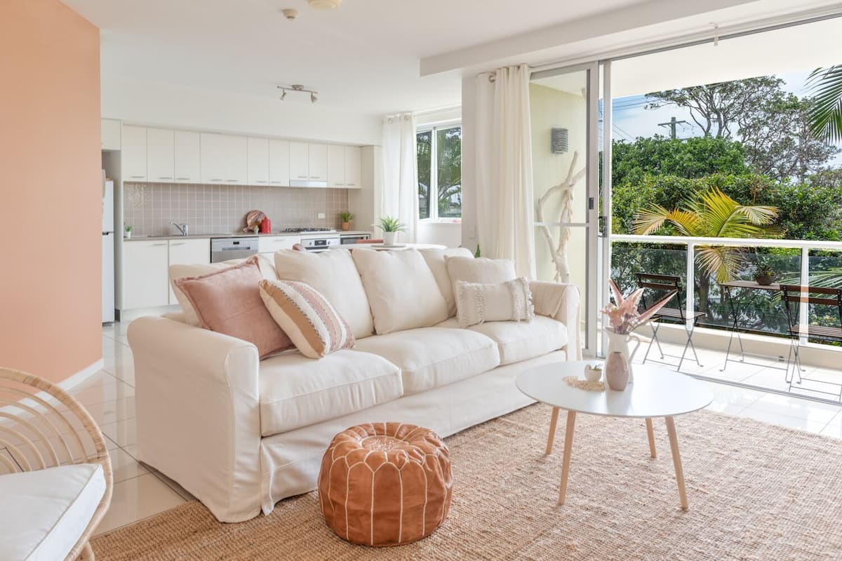 A modern living room with a white sofa, a round coffee table, and a brown ottoman. A kitchen with white cabinets is visible through an open door.