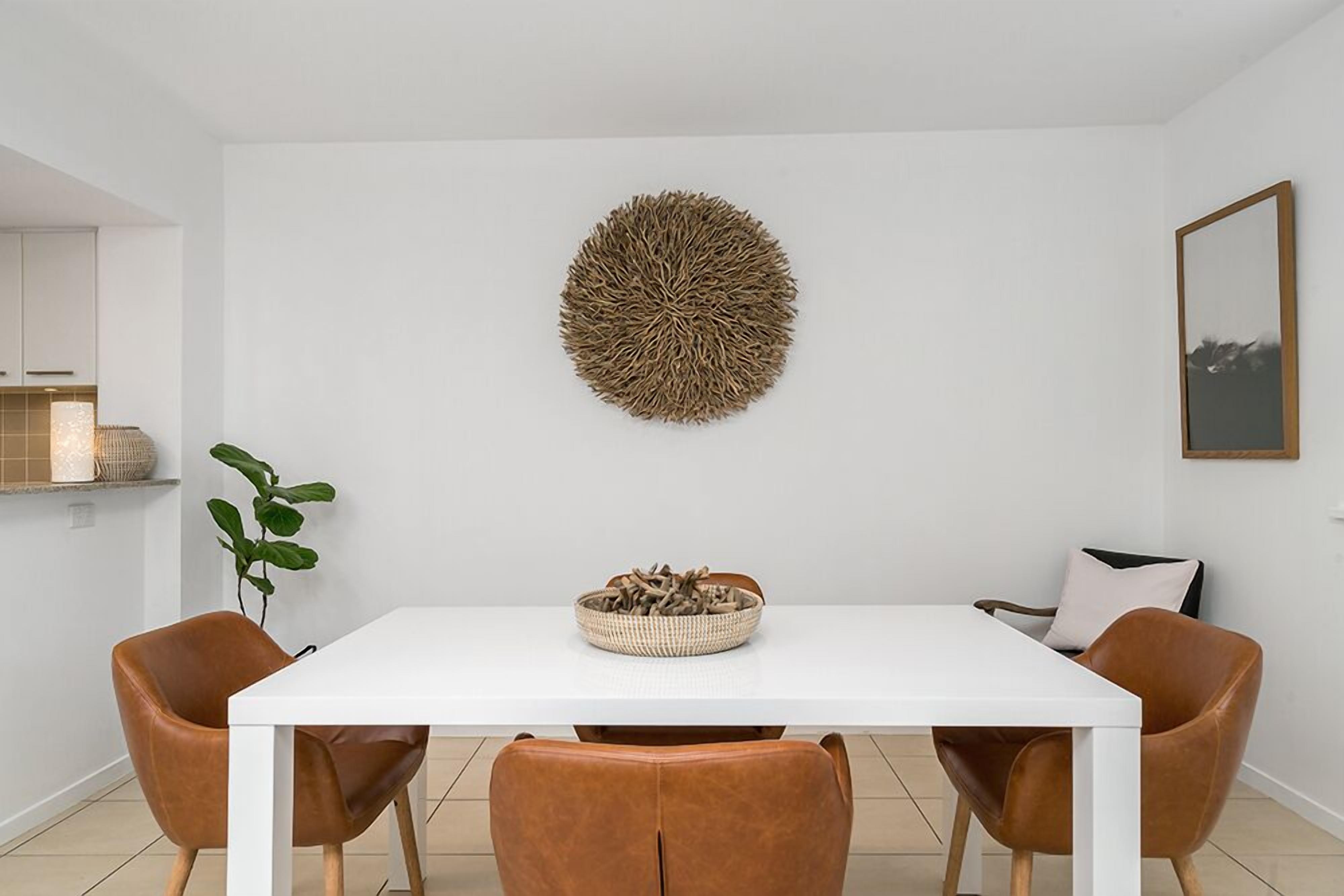 Un espace repas moderne avec une table blanche, quatre chaises marron, une œuvre d’art murale en fibres tressées et une plante.