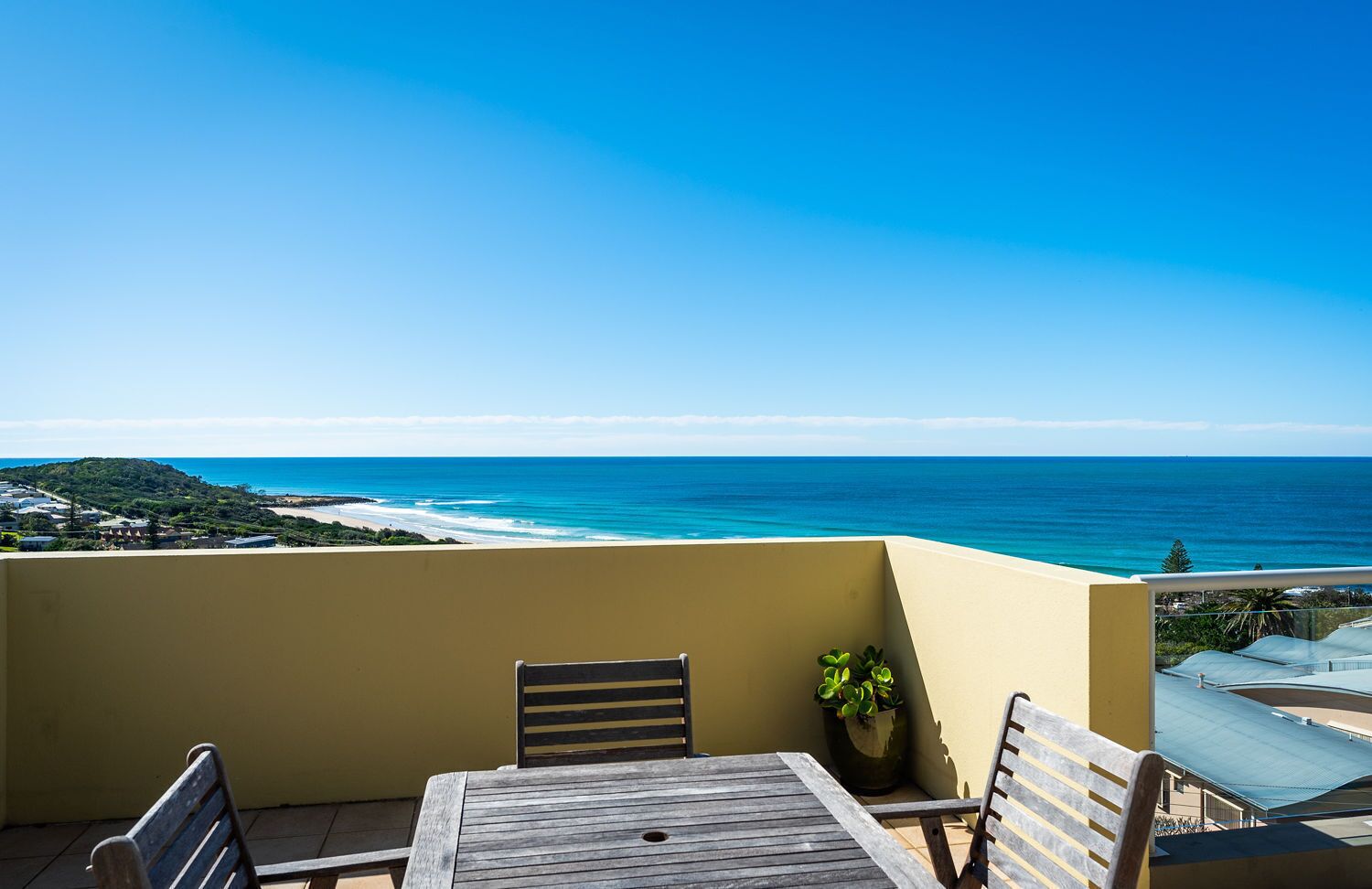 A balcony with a wooden table and chairs, overlooking a beach and ocean.