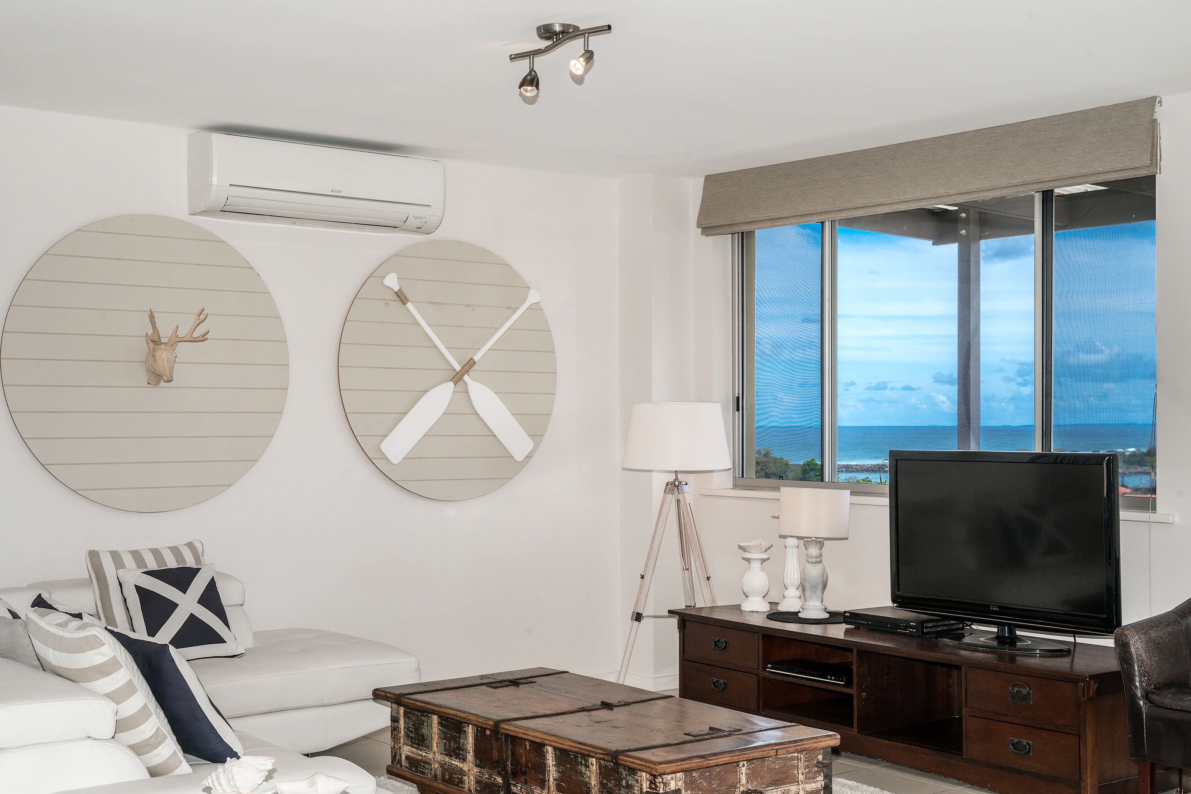 A modern living room with a white sofa, a wooden coffee table, a TV on a wooden stand, and a large window with a view of the ocean.
