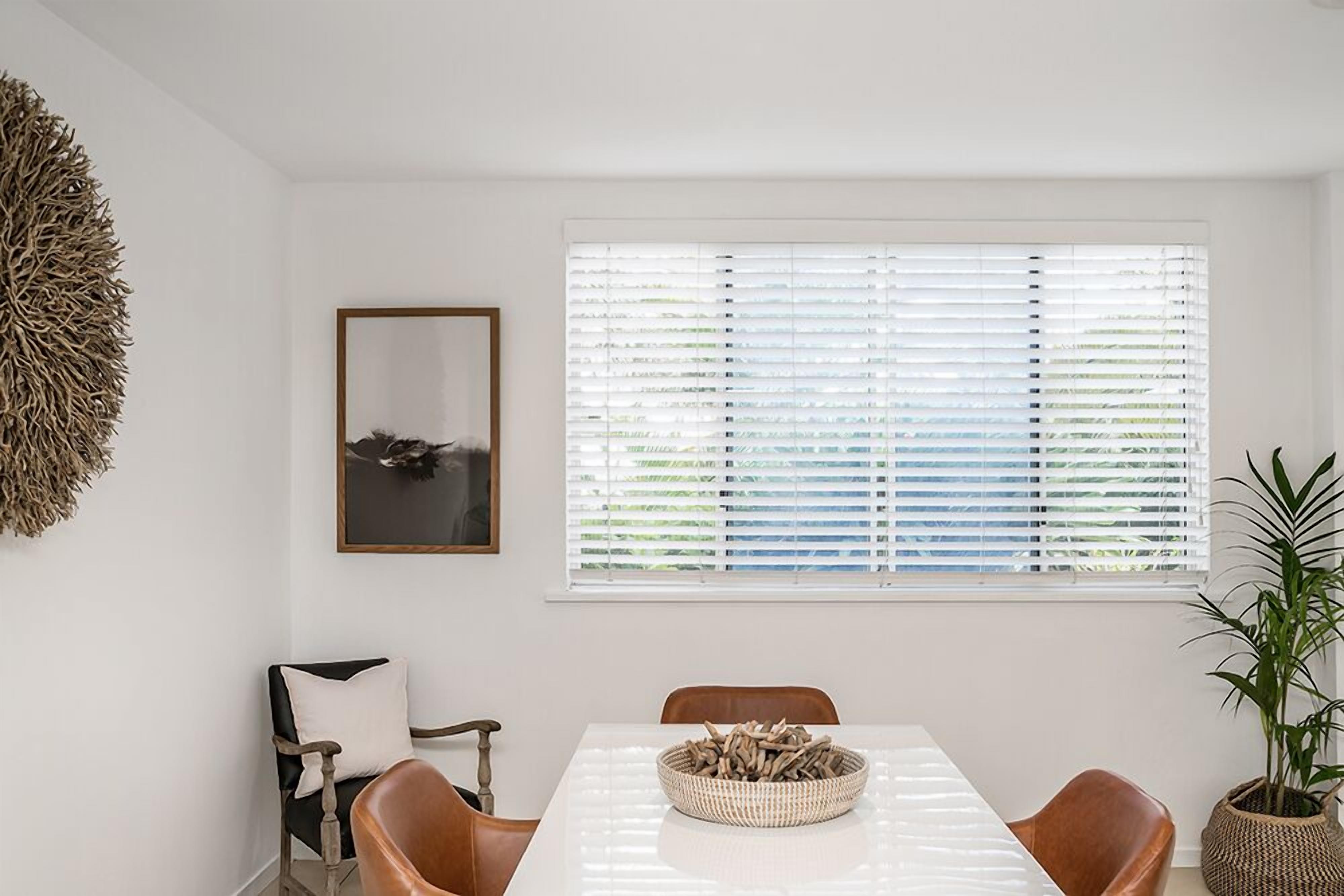 A dining area with a white table, two brown chairs, a framed picture on the wall, and a large window with blinds.