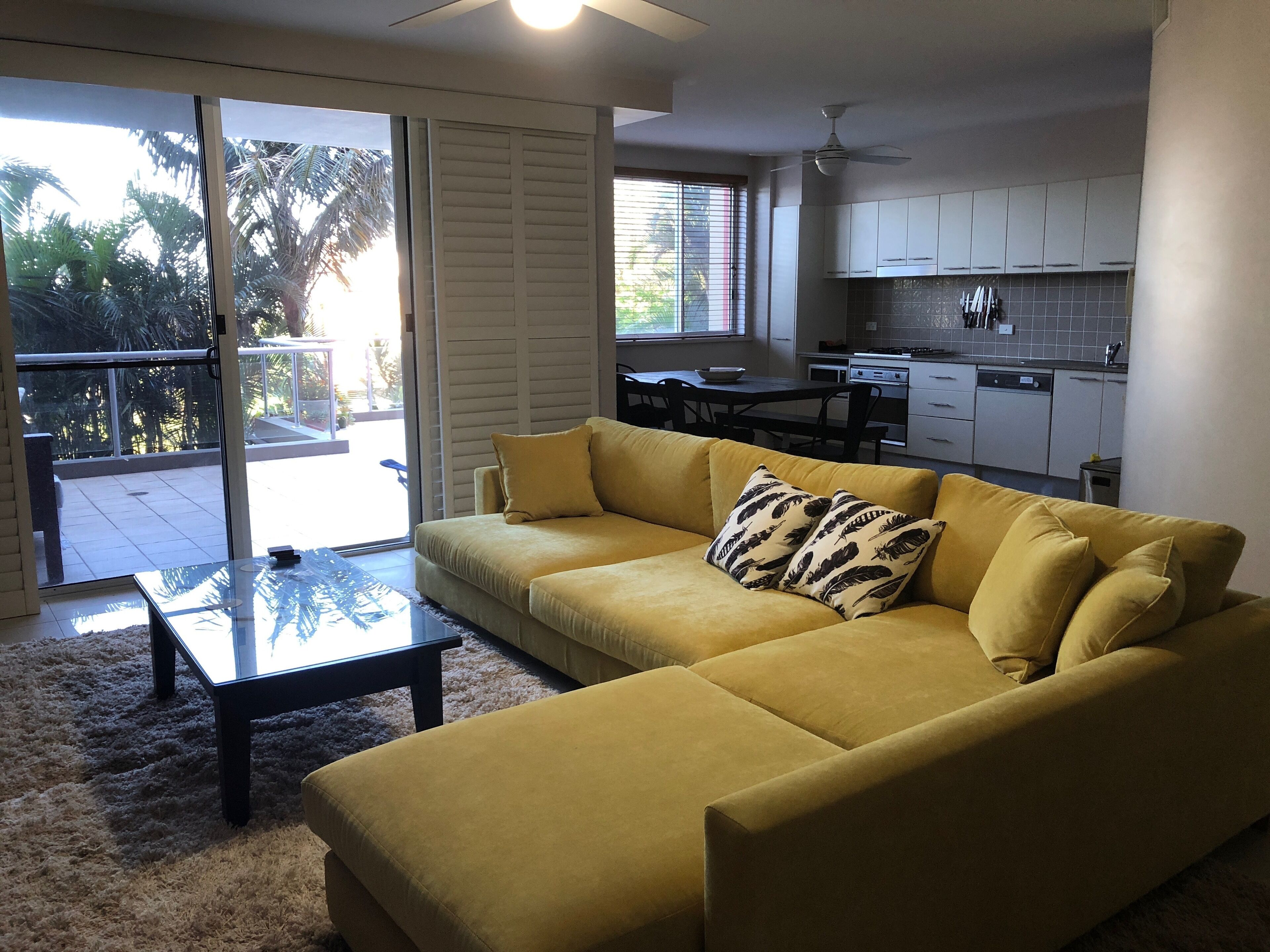 A living room with a yellow sofa, a glass coffee table, and a view of a balcony with palm trees.