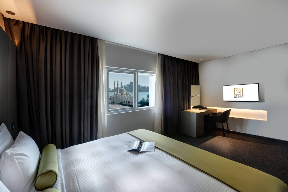 A hotel room with a bed, a desk, a chair, a TV, and a view of a cityscape.
